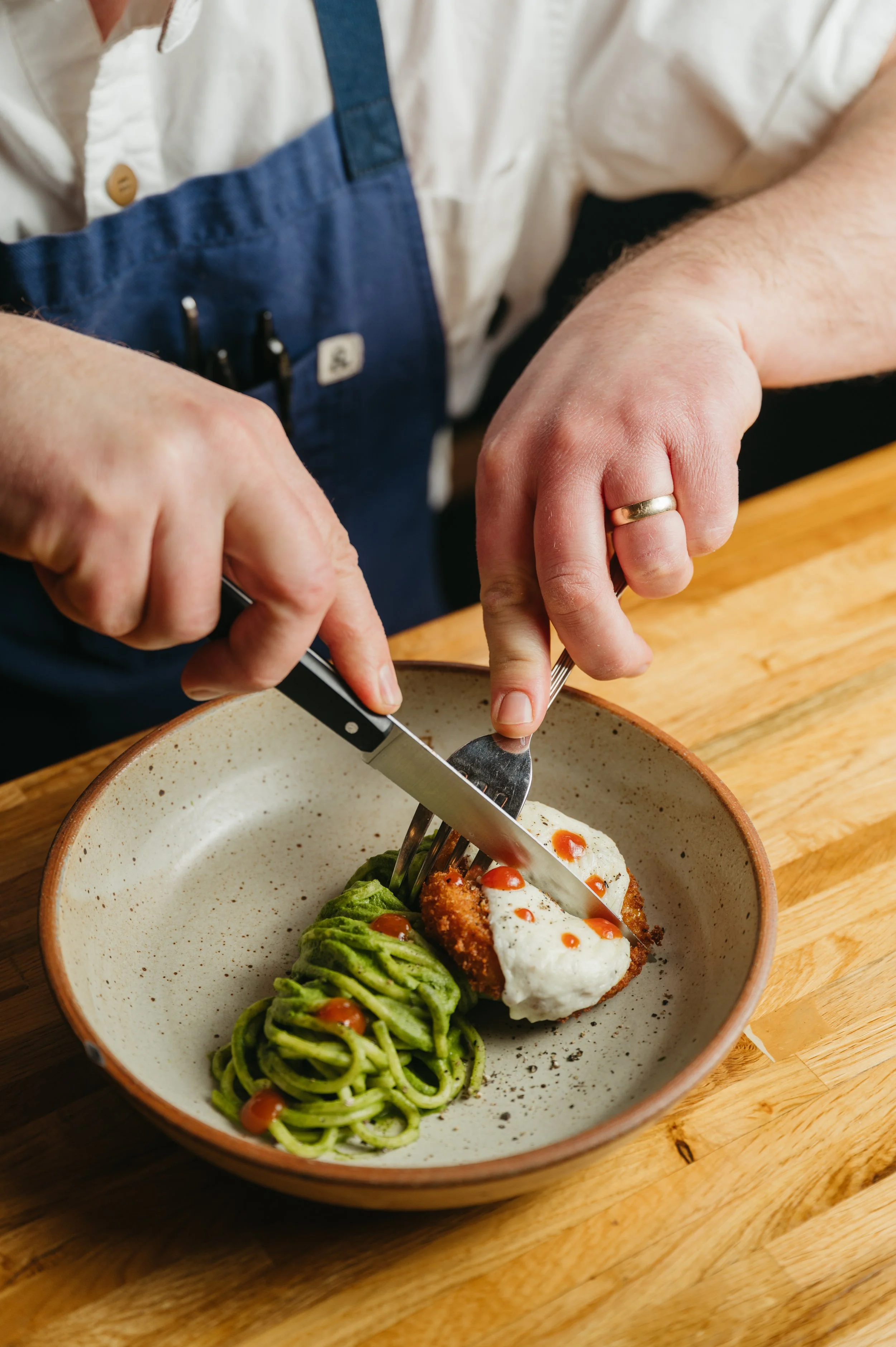 Person cutting into a dish of fried fish topped with sauce and a poached egg, served with pesto pasta, on a wooden table.