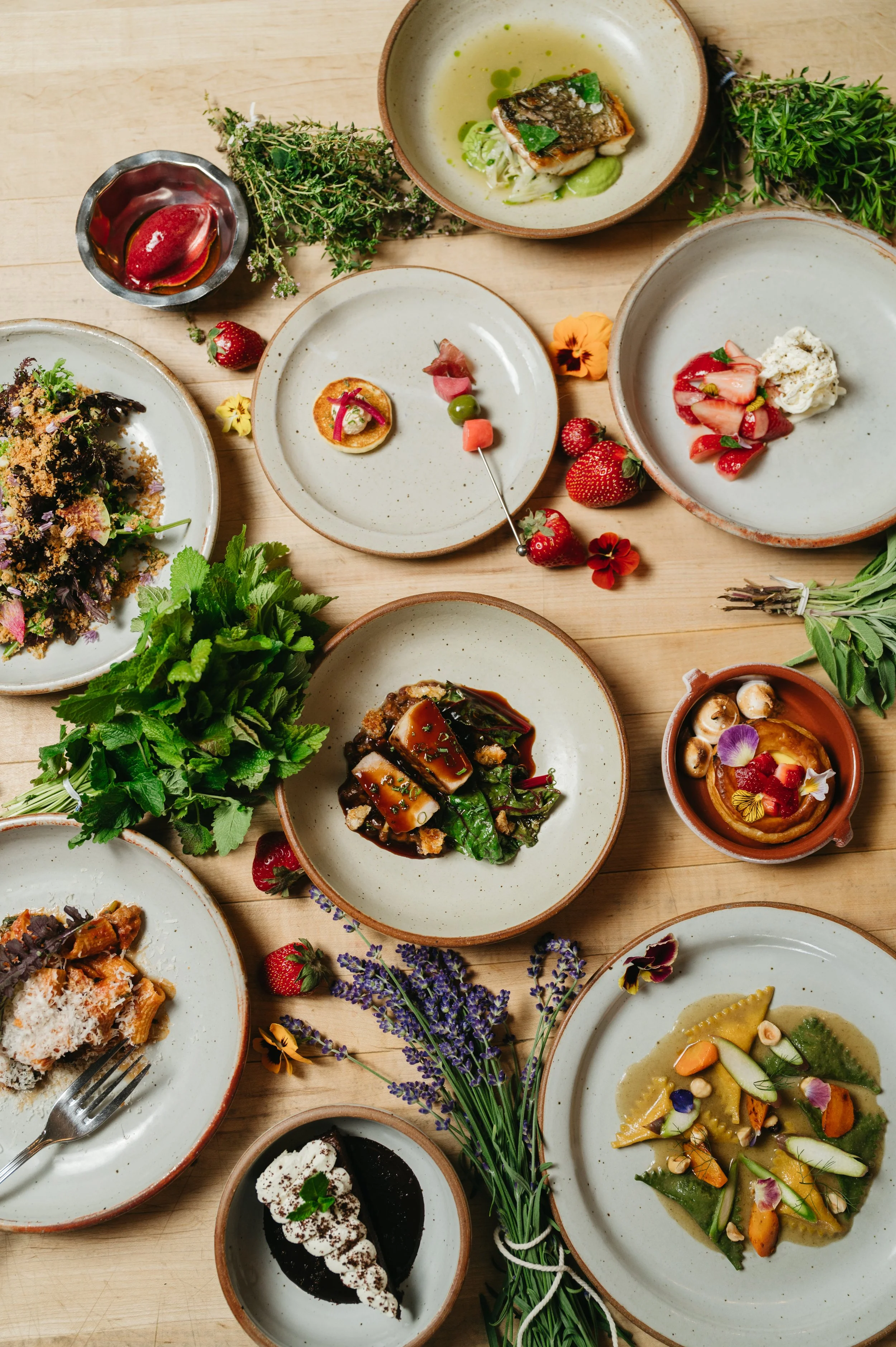A variety of gourmet dishes on a wooden table, including pasta, salads, and desserts, garnished with fresh herbs and edible flowers.