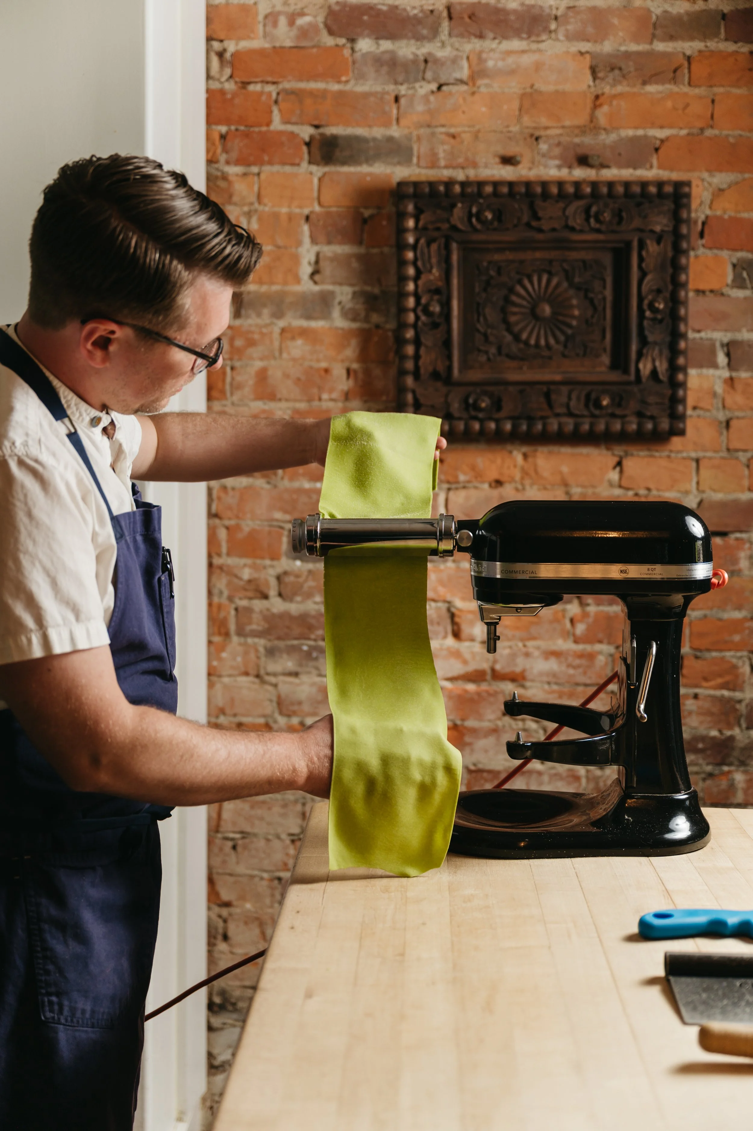A man wearing glasses and a blue apron, standing at a wooden workbench, is using a pasta roller machine to flatten green dough. The background features a brick wall with a decorative framed panel.