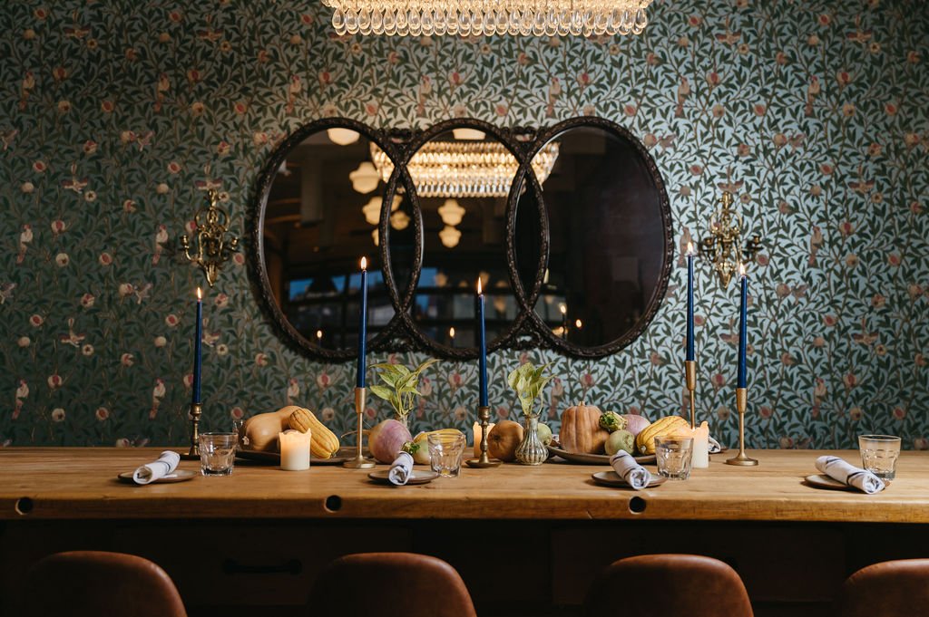 A rustic dining table with candles, vegetables, and napkins, set in a room with patterned wallpaper, mirrors, and chandeliers.
