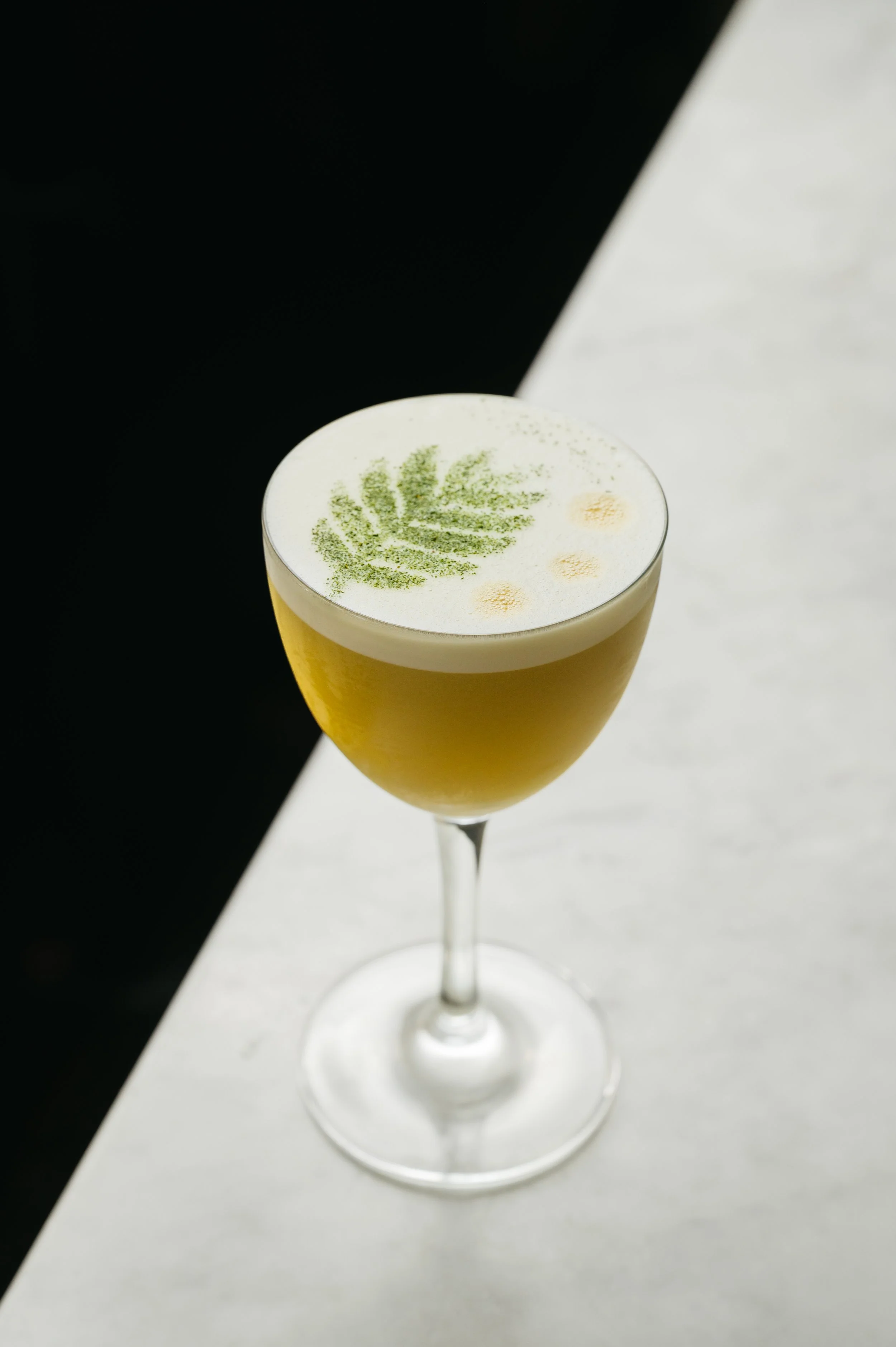 A cocktail in a tall, elegant glass with a frothy white top, garnished with a green leaf pattern and small yellow circles, set against a split black and white background.