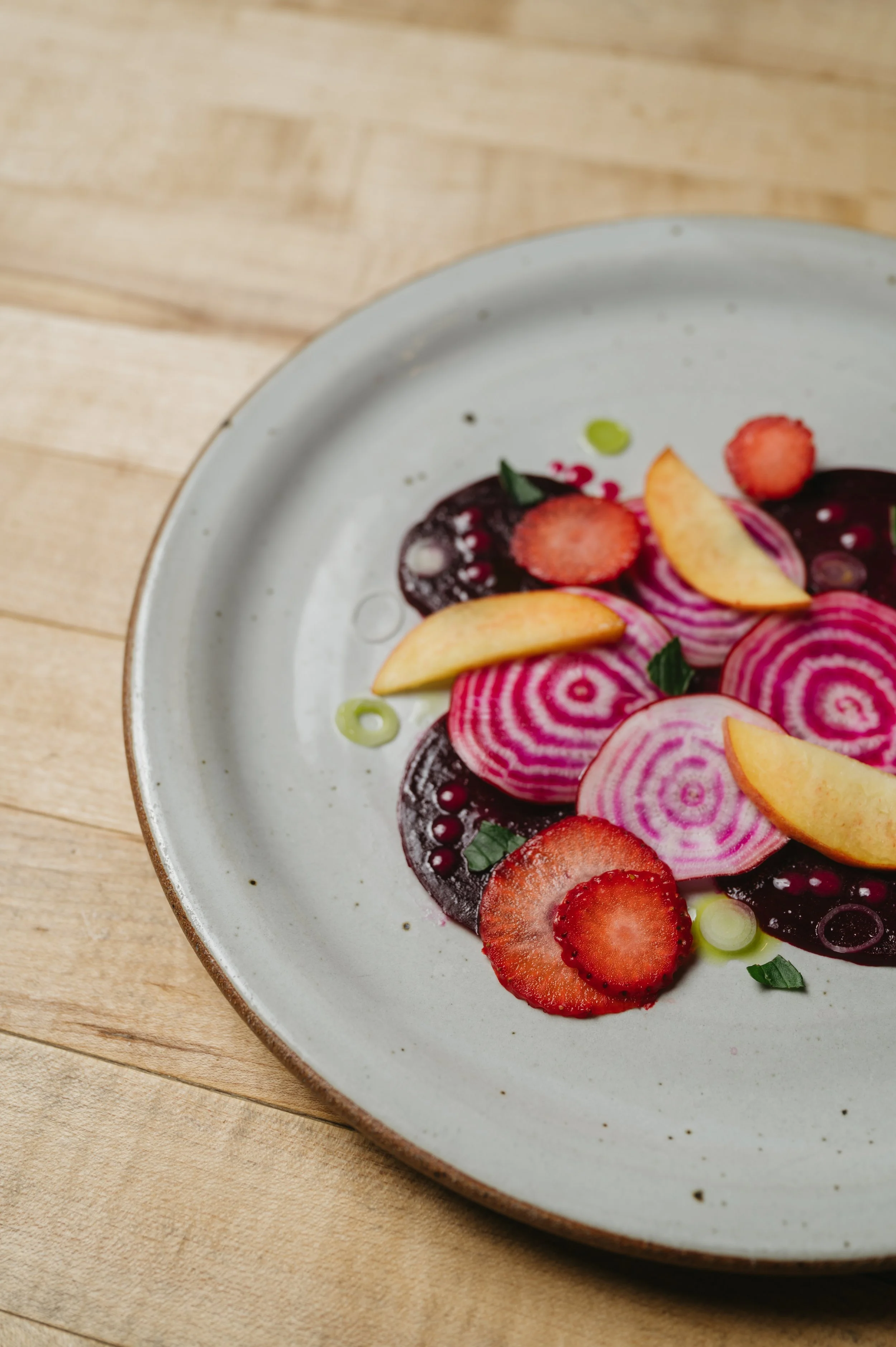 Close-up of a plate with sliced strawberries, radishes, and peach slices, garnished with green onions, on a wooden table.