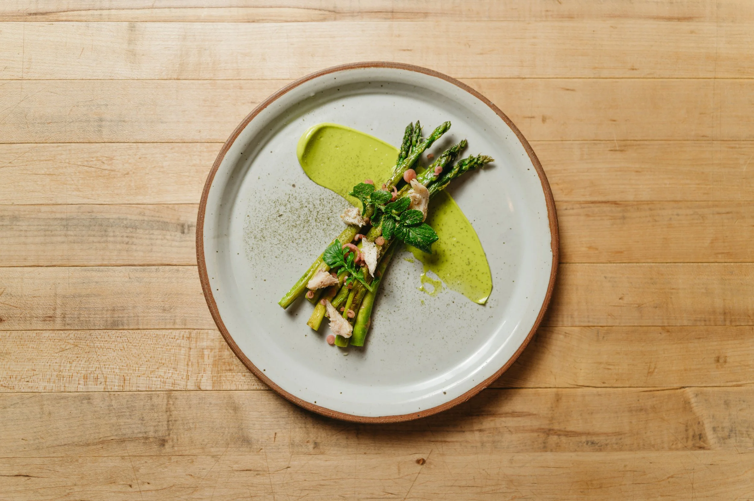 A plate of cooked asparagus spears topped with herbs and pink peppercorns, with a drizzle of green sauce on a white plate with a brown rim, on a wooden table.