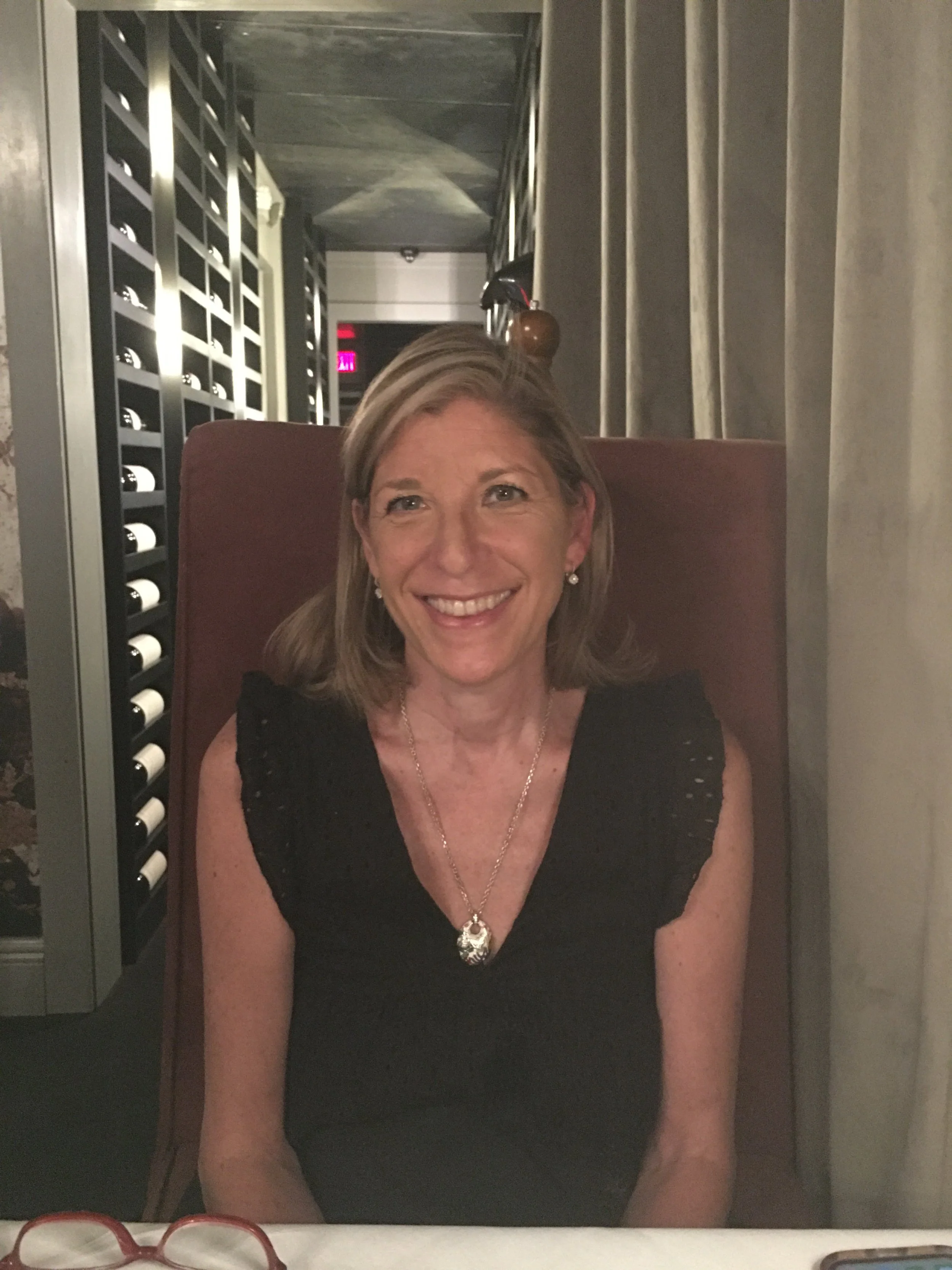 A smiling woman with shoulder-length blonde hair wearing a black dress with pearl earrings and a necklace, sitting at a table near a wine rack.