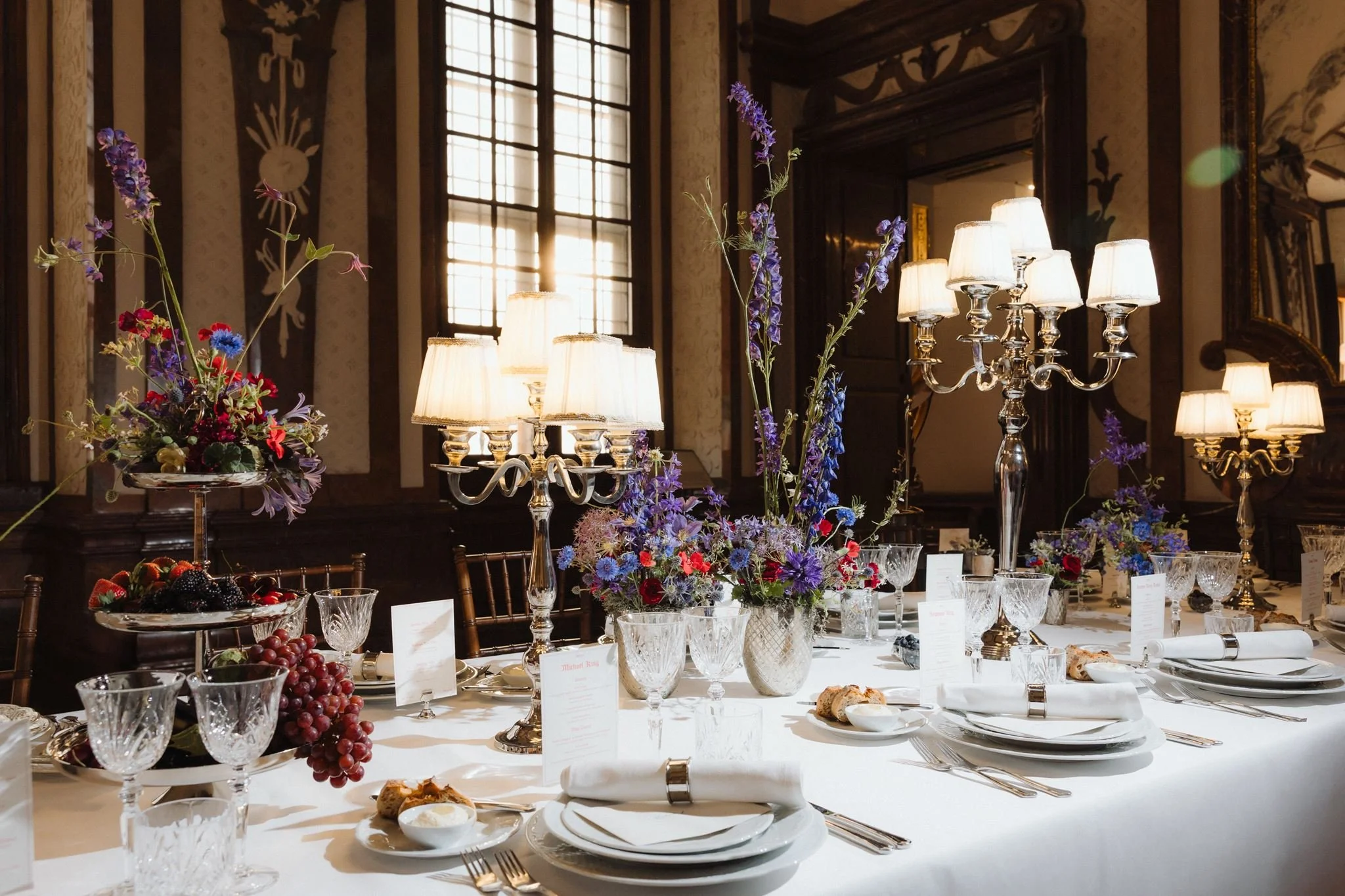 A banquet table set with crystal glassware, white plates, silverware, and cloth napkins in a large ornate room with high windows. The table is decorated with multiple floral arrangements in crystal vases and candelabras with small lamps. There is a fruit display with grapes and berries and several dishes of food. The room has dark wood paneling, decorative wall features, and large windows allowing natural light to illuminate the scene.