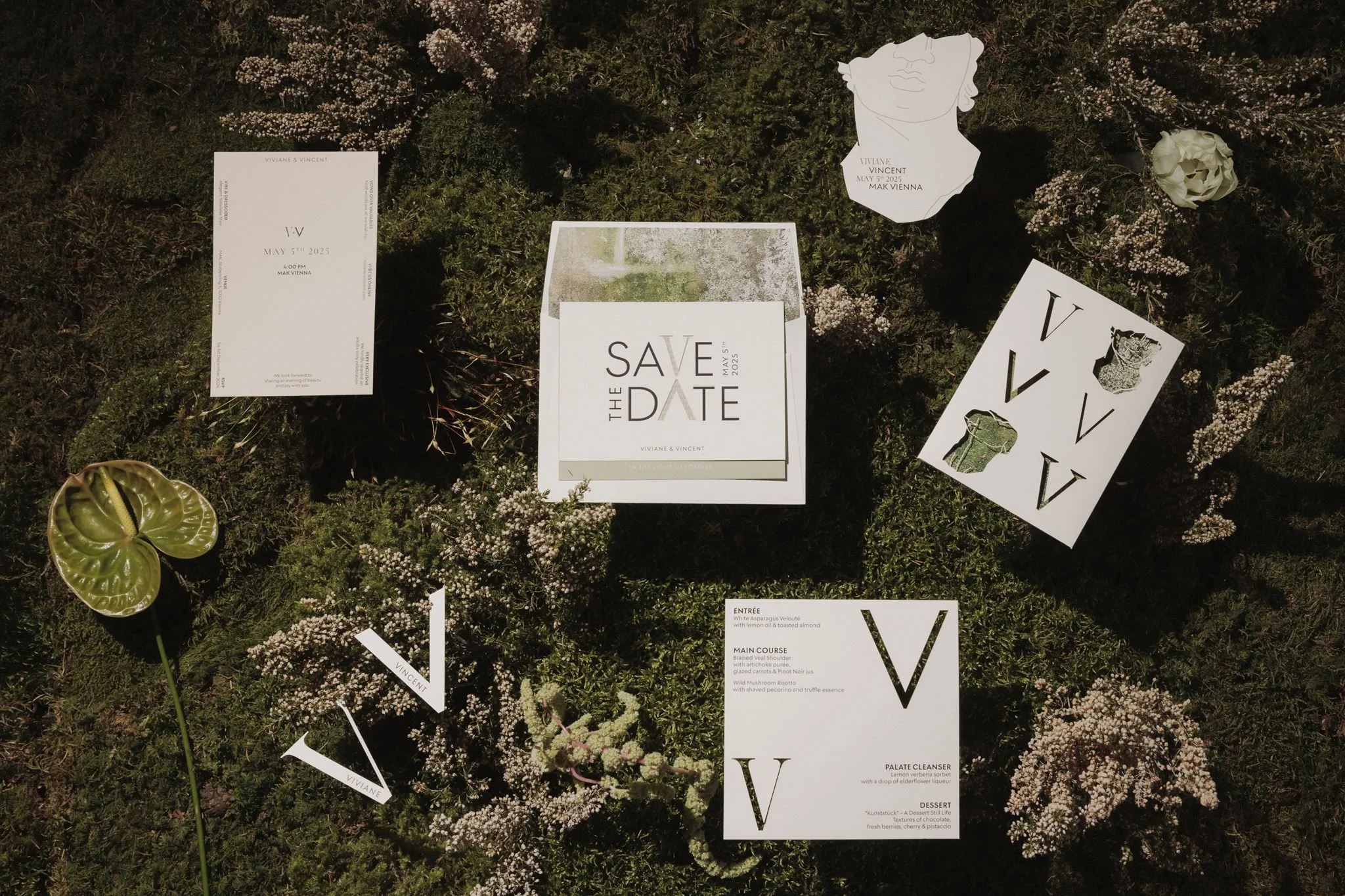 A collection of wedding or event stationery on moss and small flowers, including a save the date card, invitations, and menu cards with minimalist black and white designs.