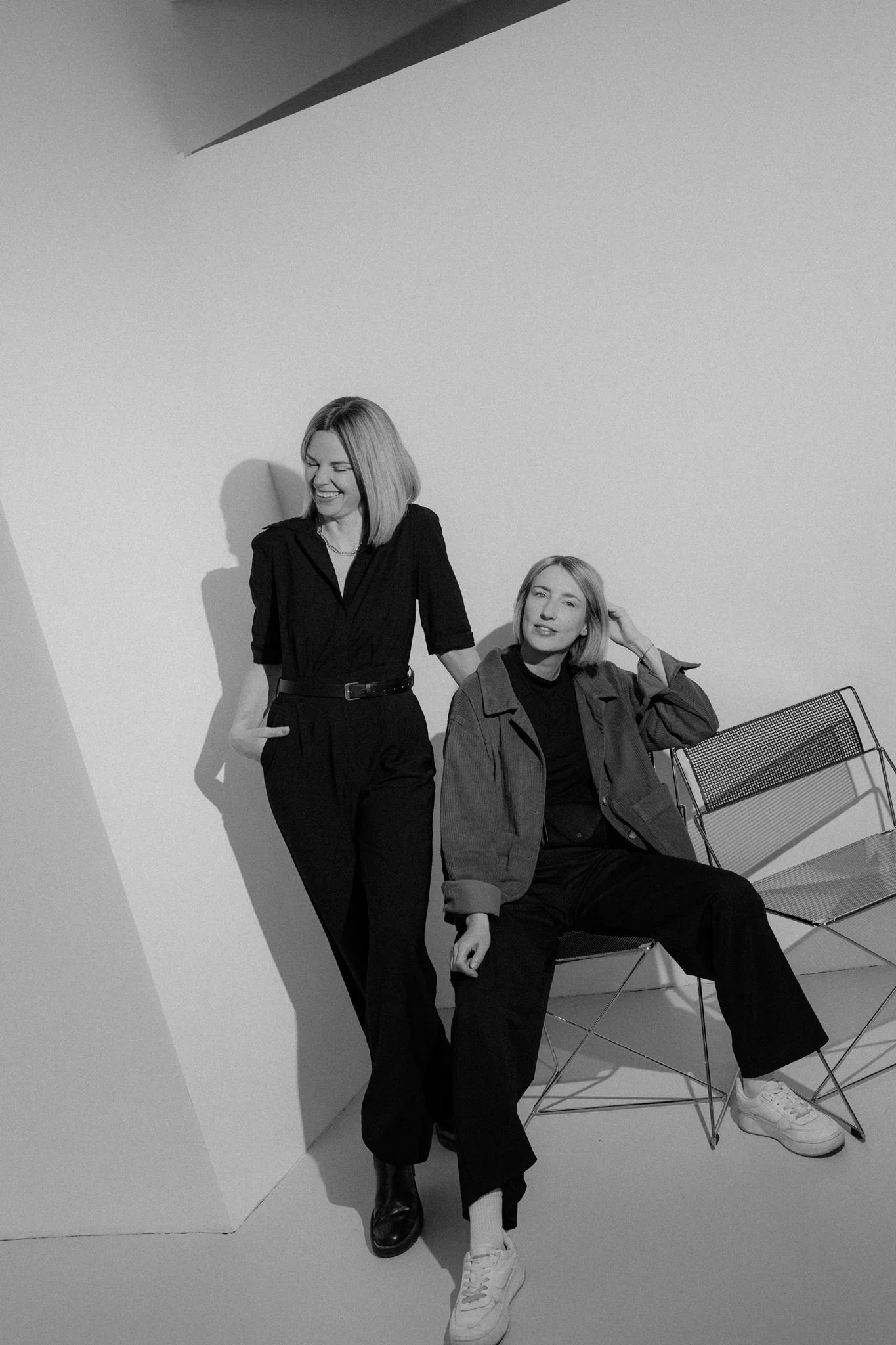 Black-and-white photo of two women in casual outfits in a studio with plain walls and a chair, smiling and relaxed.