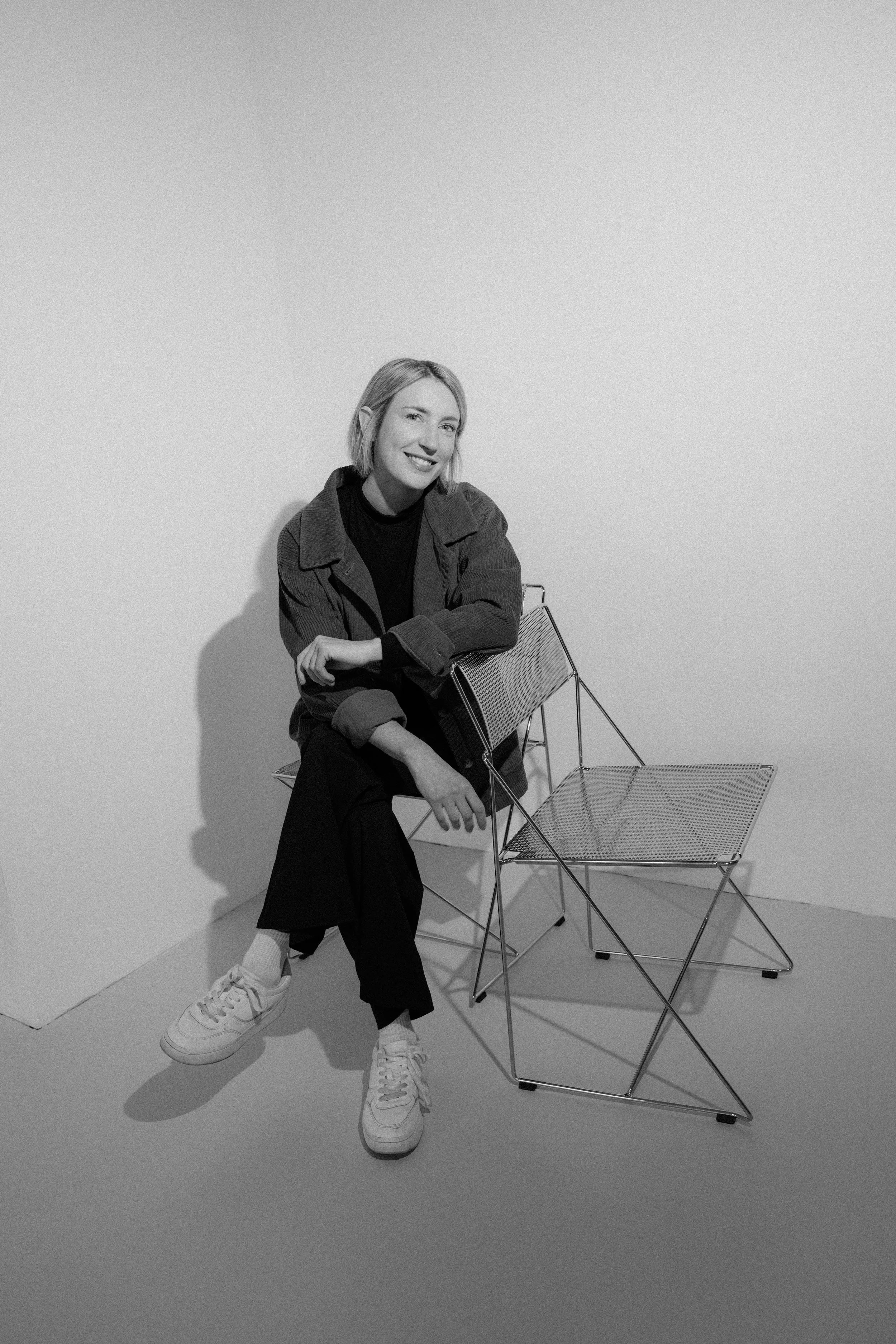 A woman with short blond hair sitting on a wire chair in a room with plain white walls. She is smiling, wearing a dark outfit with a jacket and white sneakers, with her arms crossed and one leg bent.