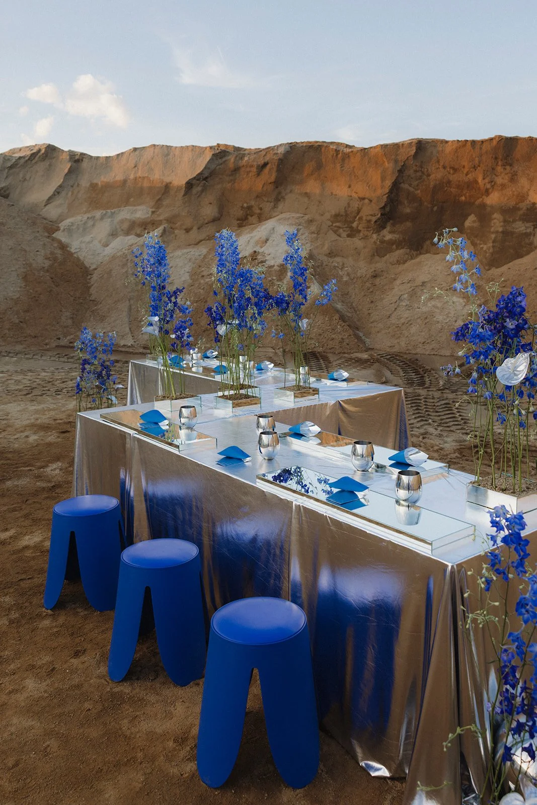 A set dining table with blue flowers and blue napkins, silver tablecloth, surrounded by blue stools, set against a desert canyon landscape at sunset.
