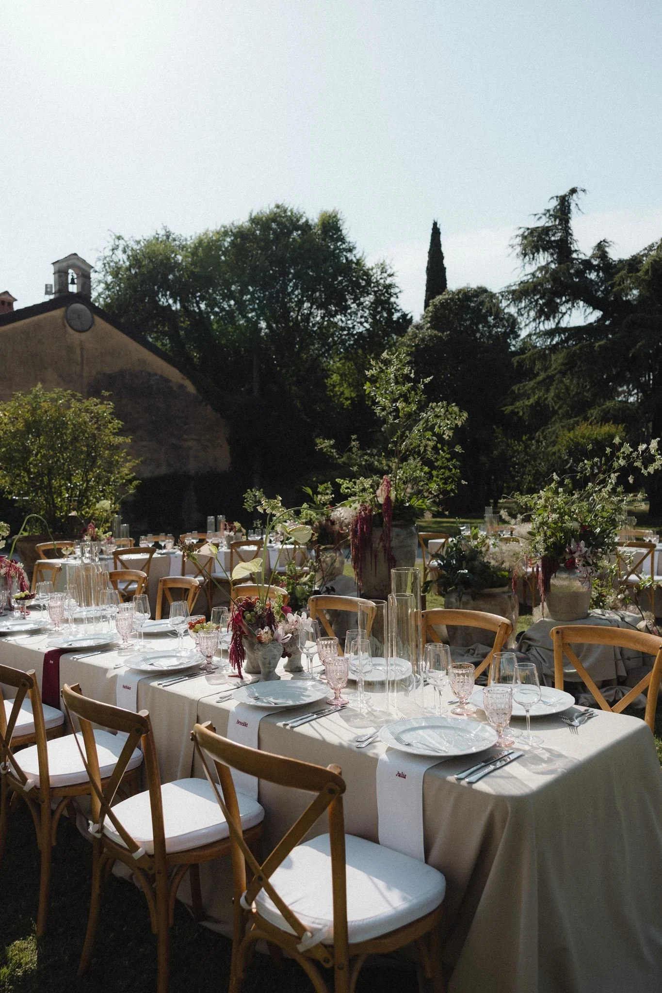 A long banquet table set outdoors with white tablecloths, pink glassware, and floral centerpieces, under a canopy of trees with a building and bell tower visible in the background.
