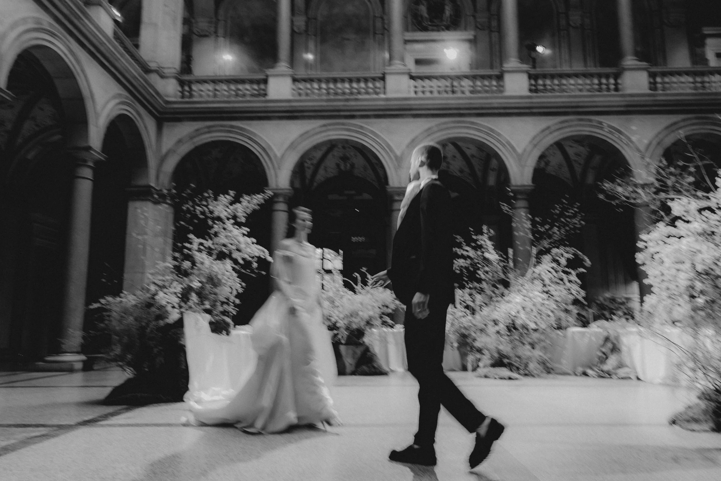 A bride and groom dance inside a decorated venue with stone arches, statues, and floral arrangements.