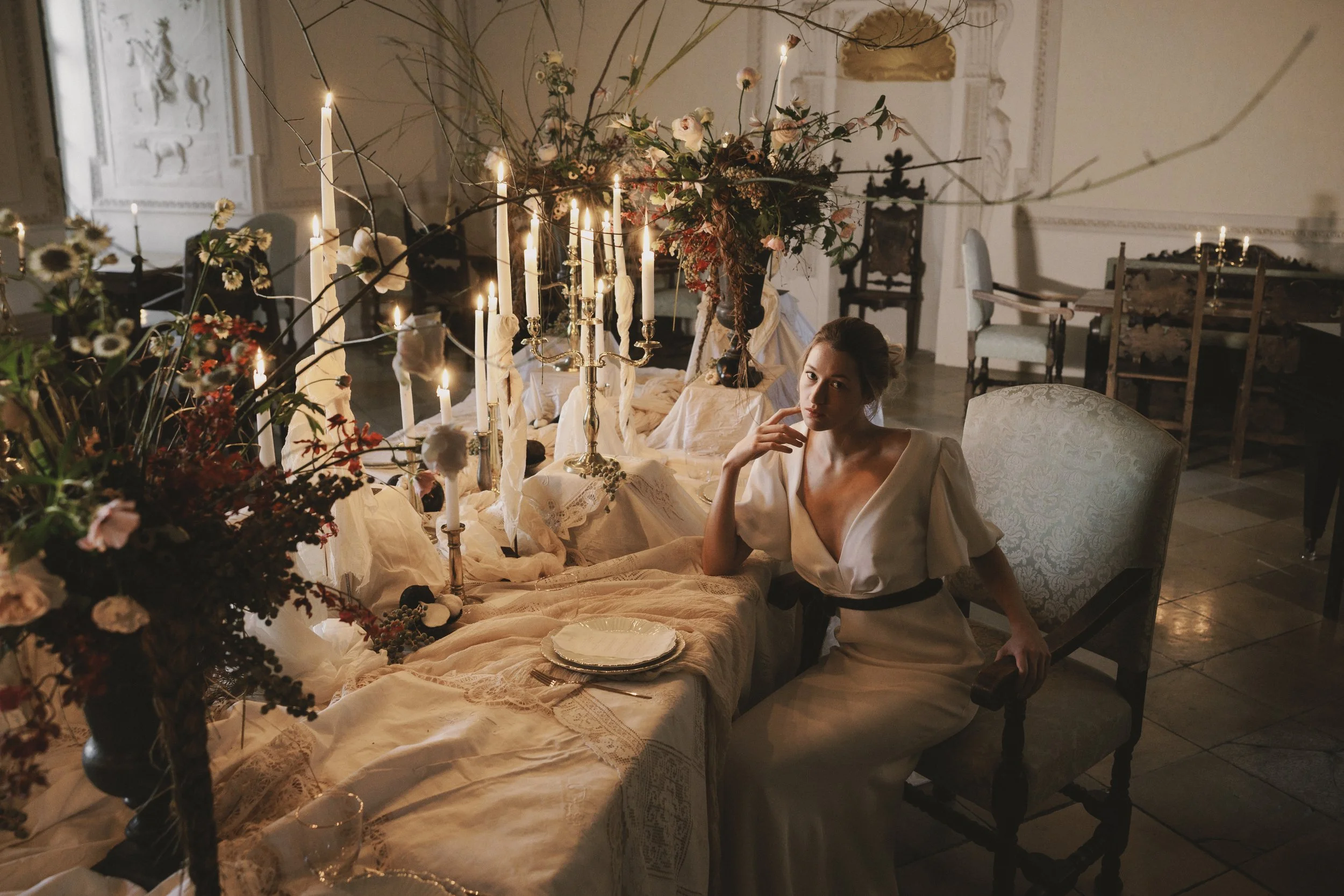 A woman in a white dress sitting at a decorated table with candles, flowers, and a large floral arrangement in a room with ornate walls and furniture.