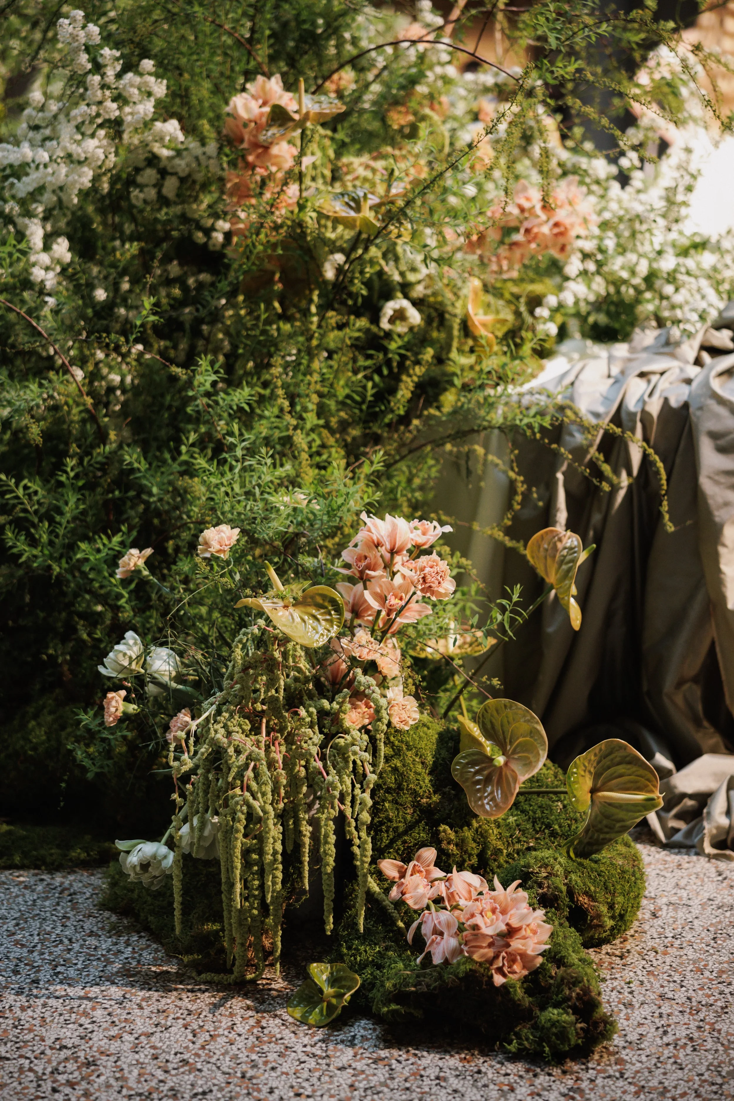 Display of various lush flowers and green plants arranged outdoors, with small rocks on the ground and a light-colored draped fabric on the right side.