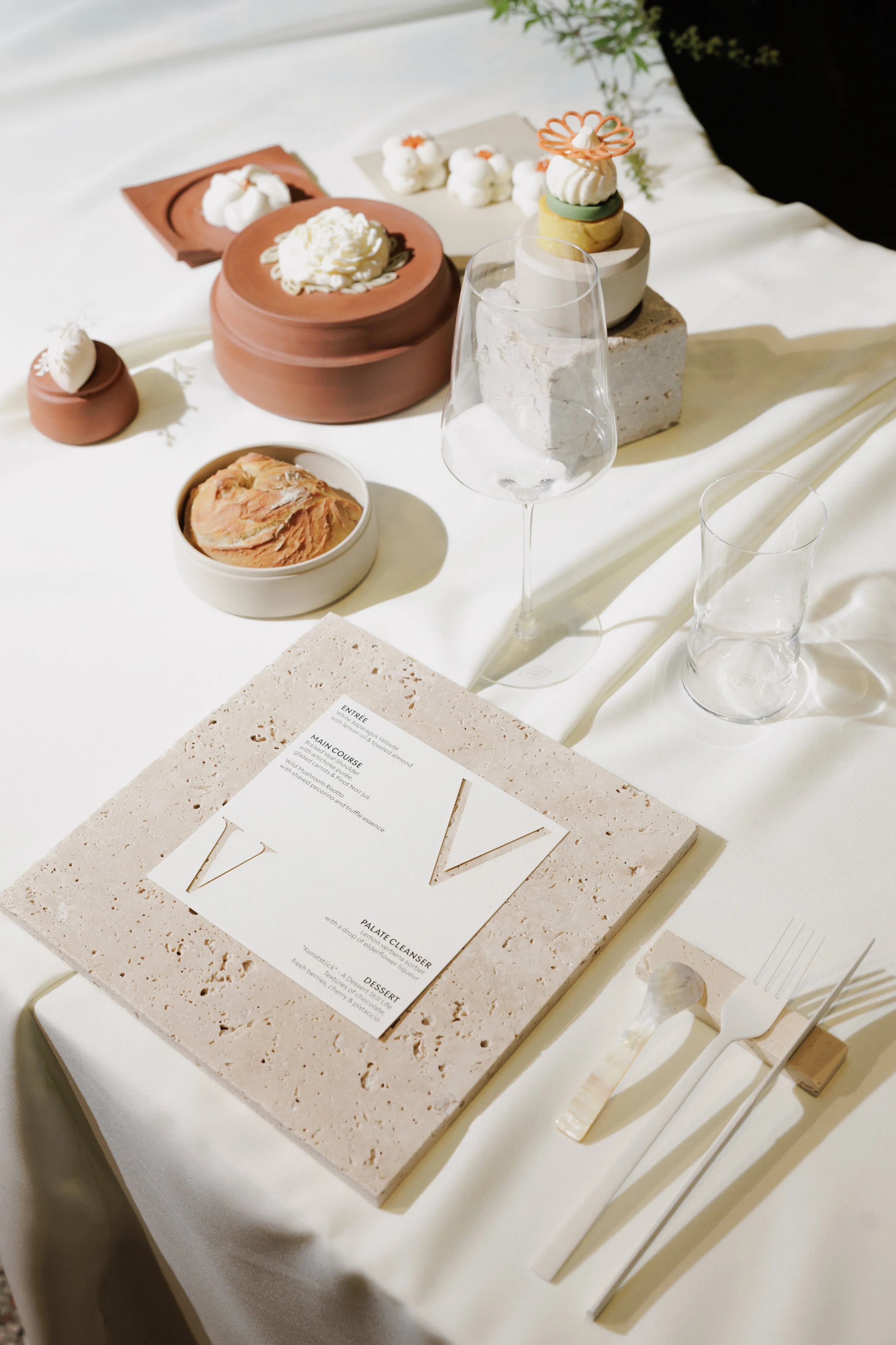 A table display featuring various dessert items, including a small chocolate mousse, a round cake topped with cream, and a layered dessert with cream and decorative toppings. There are also clear drinking glasses, a menu card, and dessert utensils on the table.