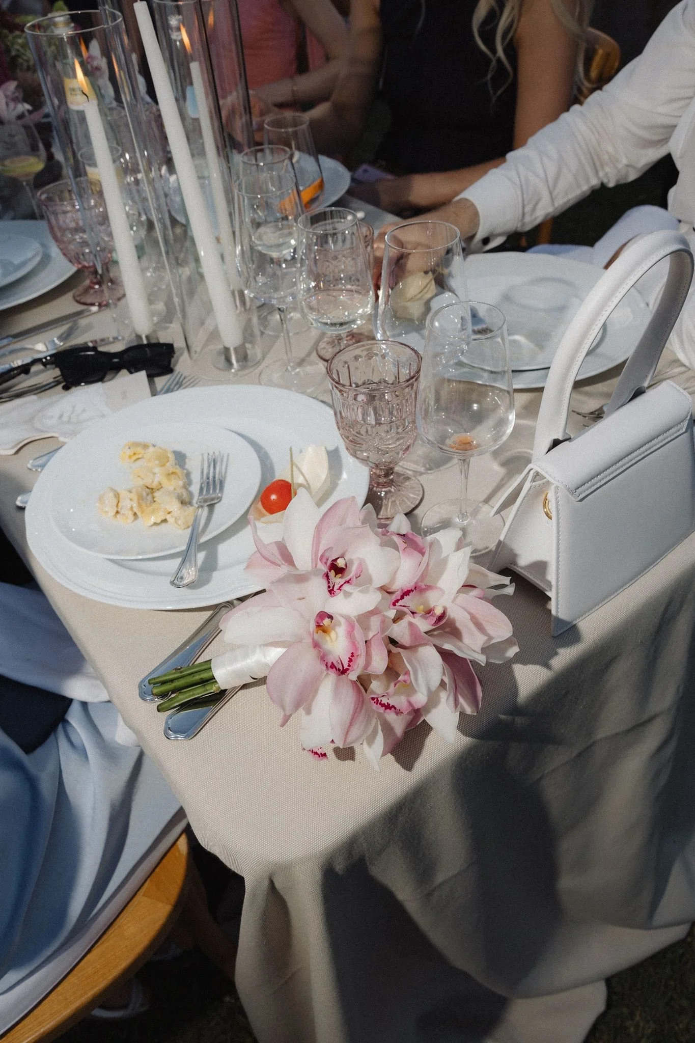 A formal dining table set with empty wine glasses, pink flowers, and a plate with cheese and cherry tomatoes. Multiple tall candles in glass holders are on the table, and people are sitting around it, dressed in formal attire.