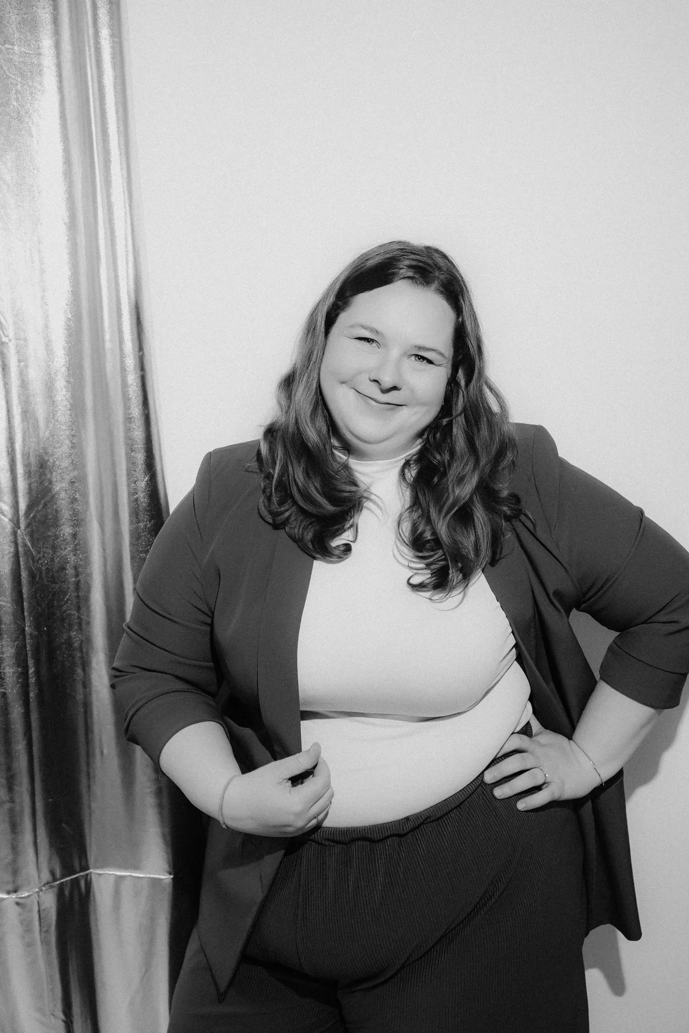 A confident woman with long wavy hair, wearing a blazer over a t-shirt, posing with hands on hips in front of a plain wall and curtain, smiling.