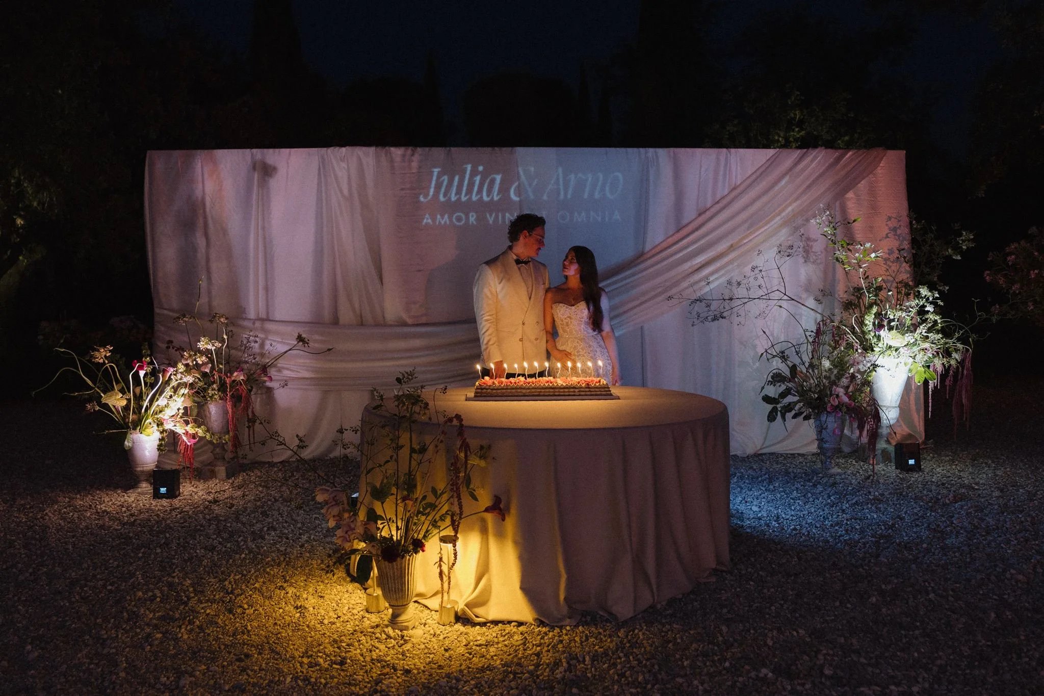 A wedding reception scene at night with a bride and groom standing together behind a decorated table with a large cake. The background features a white draped backdrop with the names Julia and Arno projected on it, and floral arrangements surround the scene.