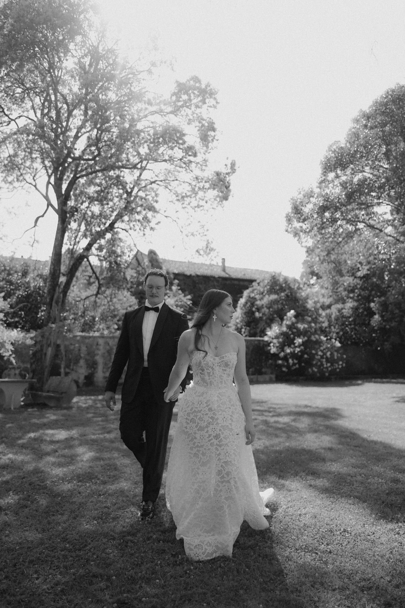 A black and white photo of a bride and groom walking outdoors on a sunny day. The bride is wearing a lace wedding gown and large earrings, and the groom is in a tuxedo with a bow tie. Trees and a house are visible in the background.