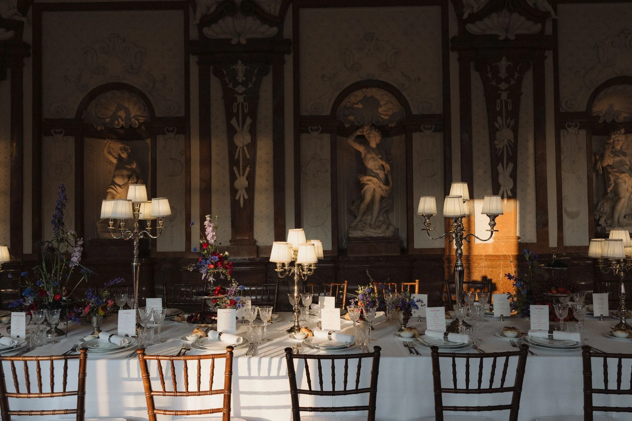 Elegant dining room with a long table set for a meal, surrounded by wooden chairs. The table has floral arrangements, candles, and place settings, with a richly decorated wall featuring statues and ornate woodwork in the background.