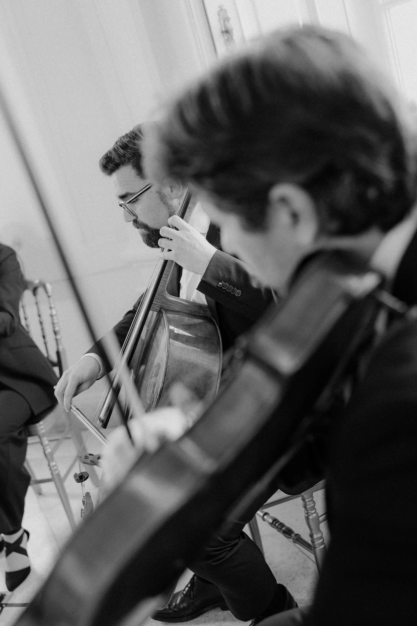 A black and white photo of a man with glasses playing a cello, with a blurred person visible in the foreground and a keyboard in the background.