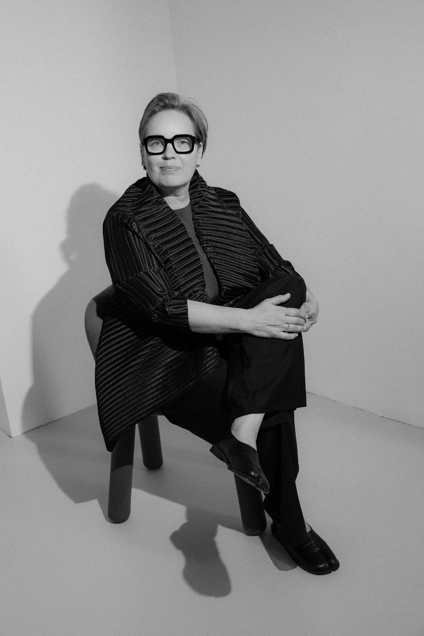 A woman sits on a stool against a plain wall, wearing large glasses, a patterned coat, and black shoes, with her hands folded on her lap.
