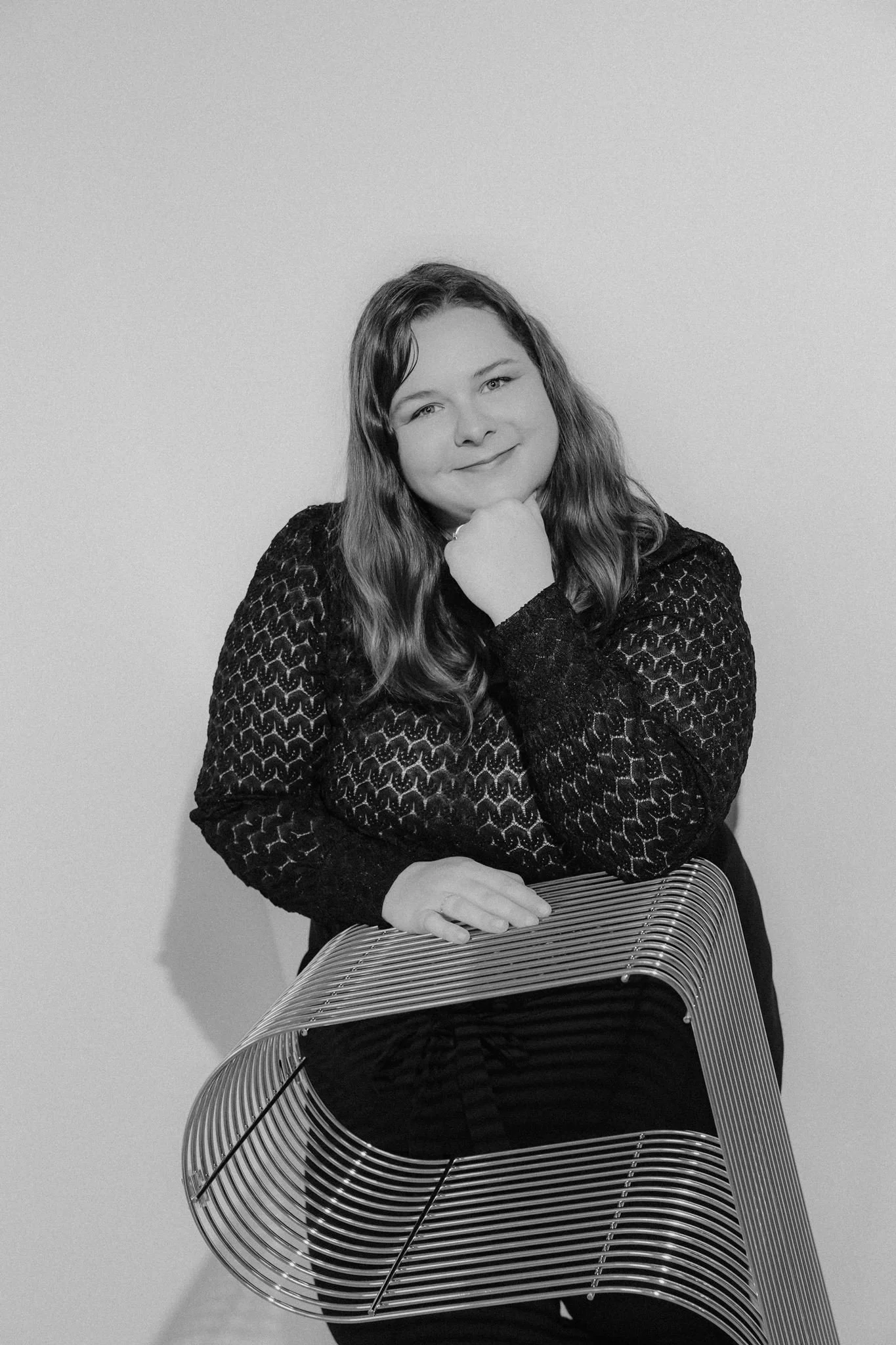 A woman with long, wavy hair wearing a patterned dark top, resting her chin on her hand, posing with a modern, curved metal chair in front of a plain wall.