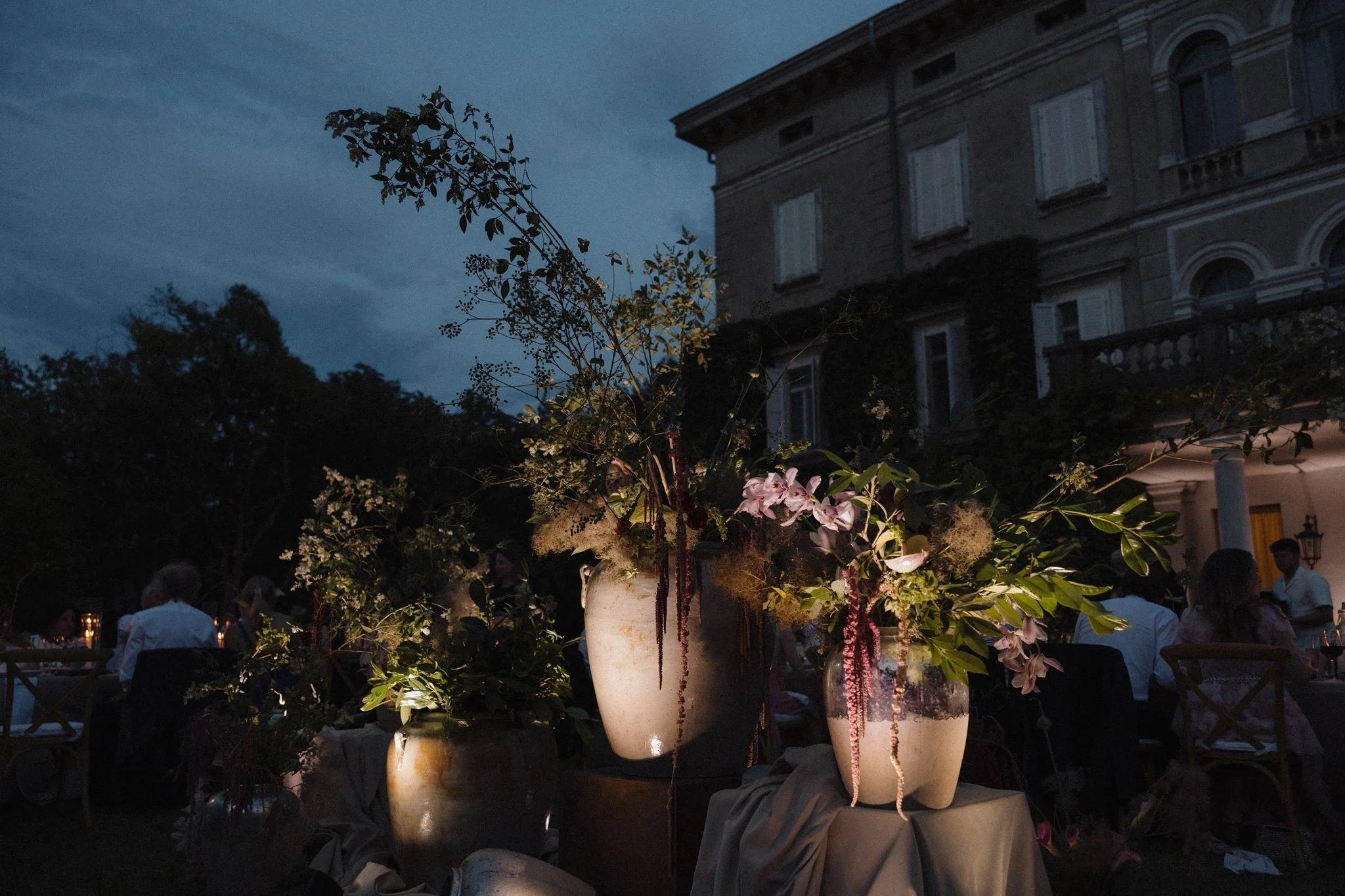 Large urns filled with pink and white flowers, illuminated at night, part of an outdoor gathering near a historic building with arched windows and shutters.