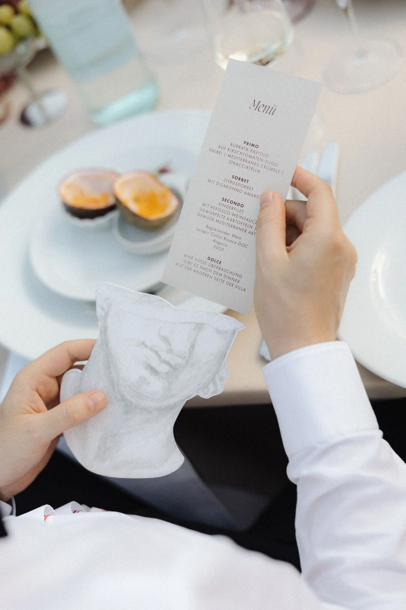 Hands holding a menu card and a paper bag over a white table with halved figs and drinks in the background.