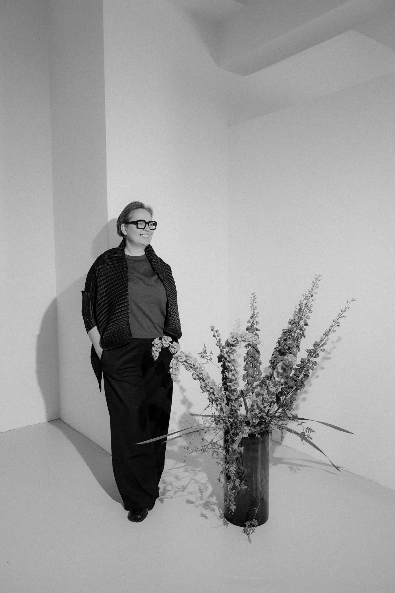 A woman with short hair, glasses, and a striped jacket standing next to a large potted plant with tall branches and flowers in a minimalist room.