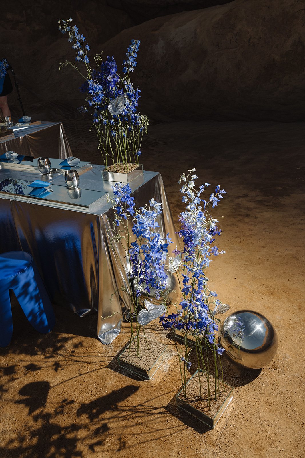 Elegant event table setup with metallic tablecloths, blue flowers in vases, and reflective decorative balls, set against a natural, rocky background.