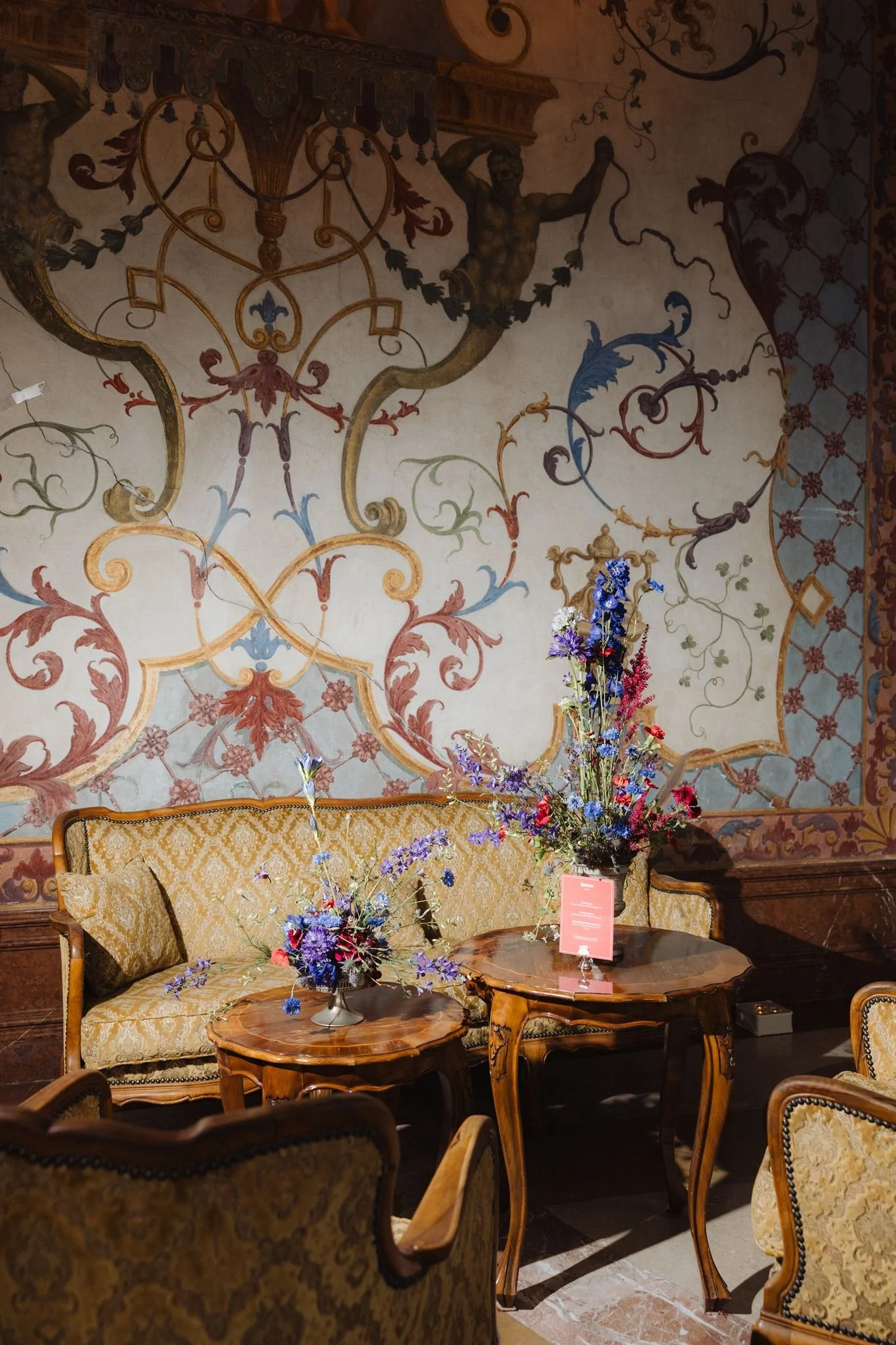 A vintage living room with ornate wallpaper and antique furniture. A floral arrangement on a wooden table is accompanied by a pink placard, and a sofa with a matching pillow is visible. The background features a decorated wall with a mural of a muscular man and intricate gold and pastel accents.