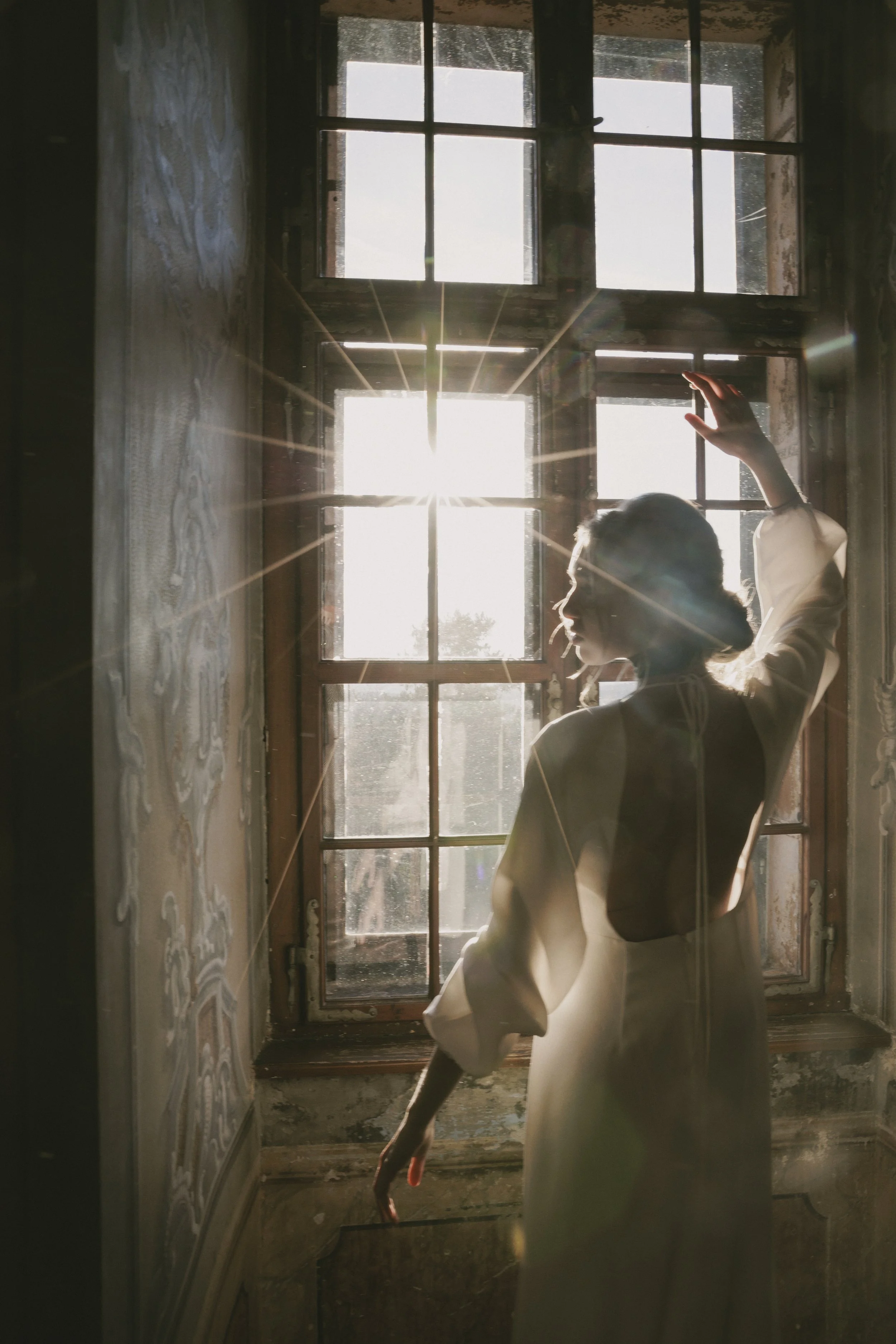 A woman with a white blazer and a dark top standing in front of a large window with sunlight streaming in. She is touching the window with her hand and looking out.