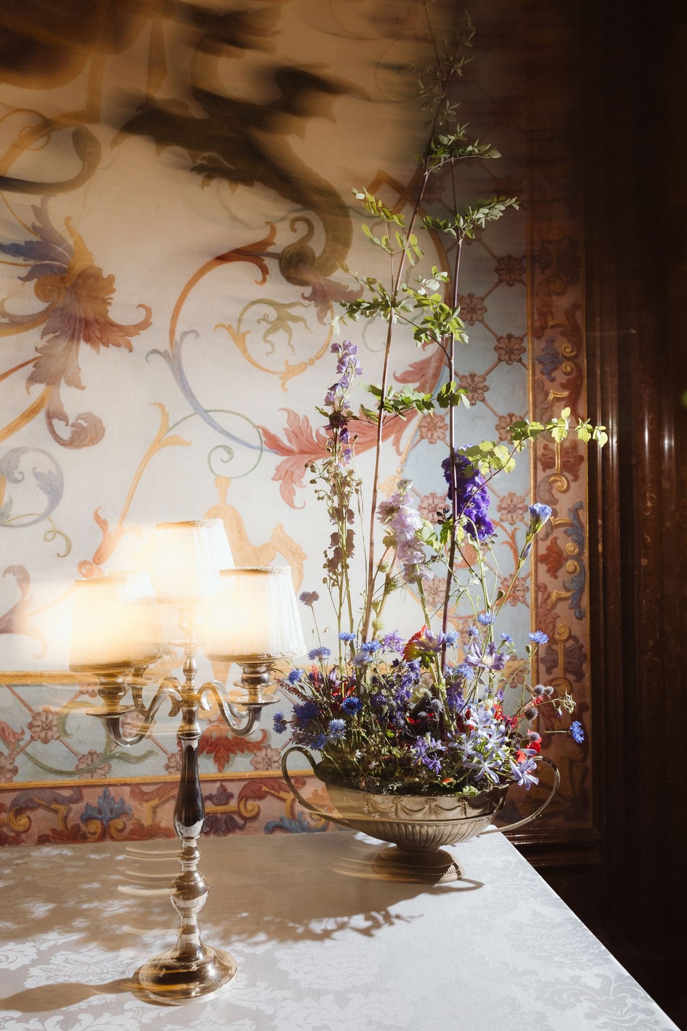 A decorative table with a silver candelabrum and a large floral arrangement in a silver bowl, in front of a wallpaper with ornate patterns in warm tones.