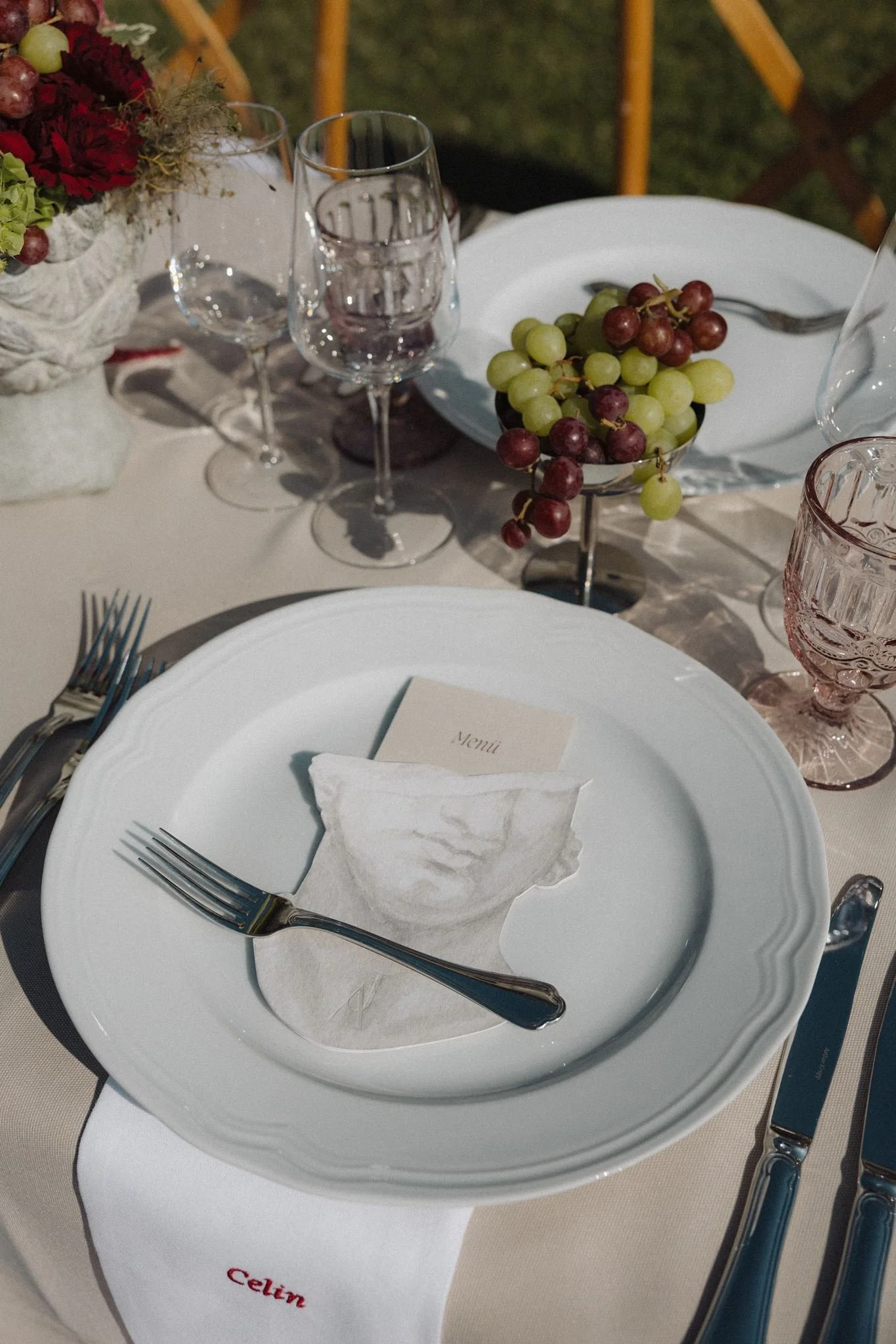 Table setting with a white plate, silver fork and knife, a glass, a folded napkin with a name tag, a bowl of green and red grapes, and a centerpiece with red flowers and green foliage, on an outdoor table with white tablecloths and a cream-colored cloth featuring red embroidered text.
