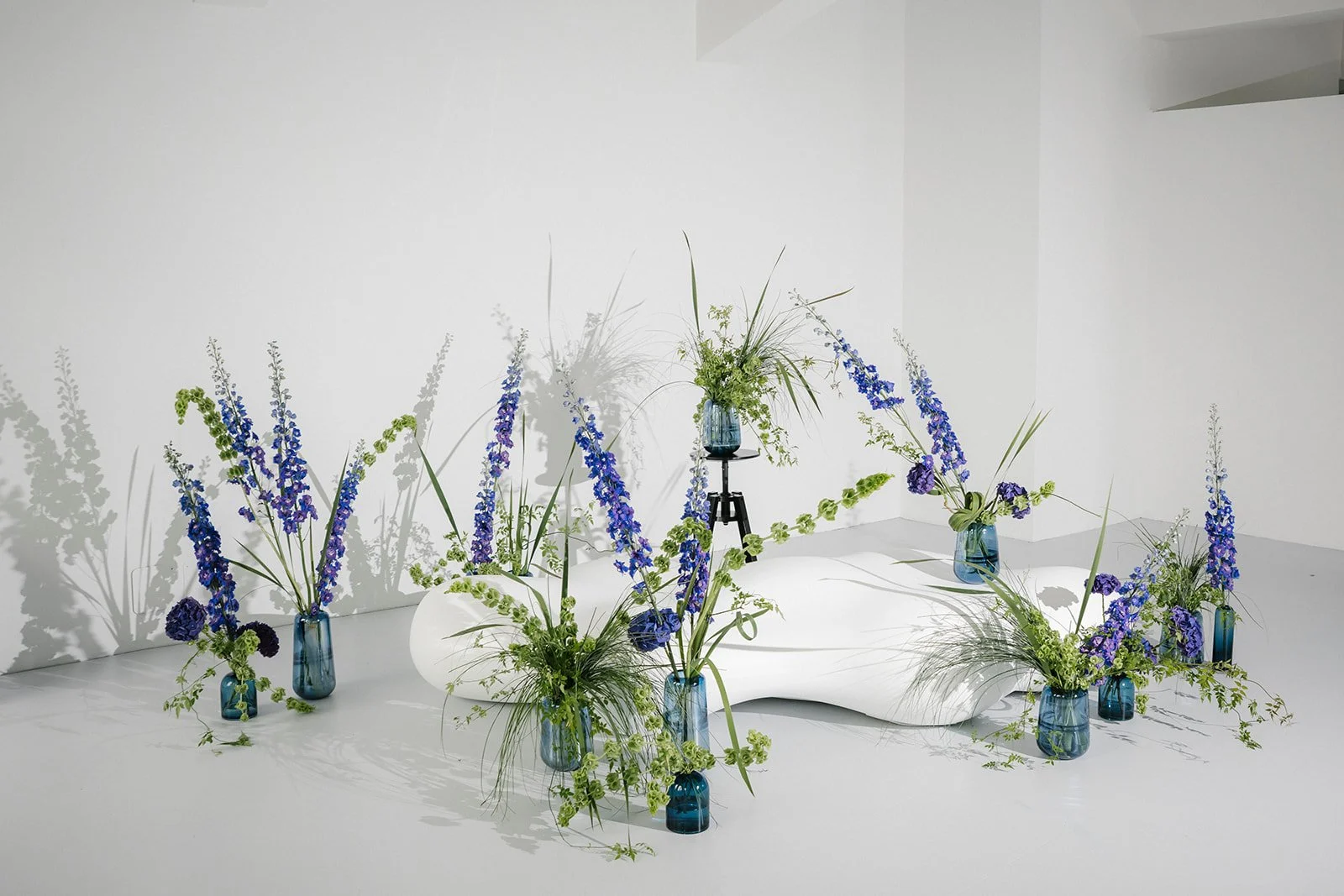 Arrangement of blue and green flowers in blue vases, with some vases on black stands, set against a white background with shadows