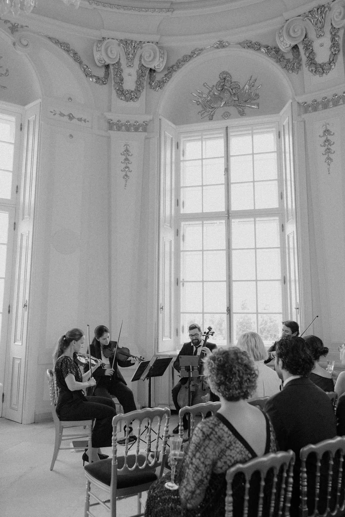 A group of musicians playing string instruments during a concert in a room with large windows and elegant architectural details.