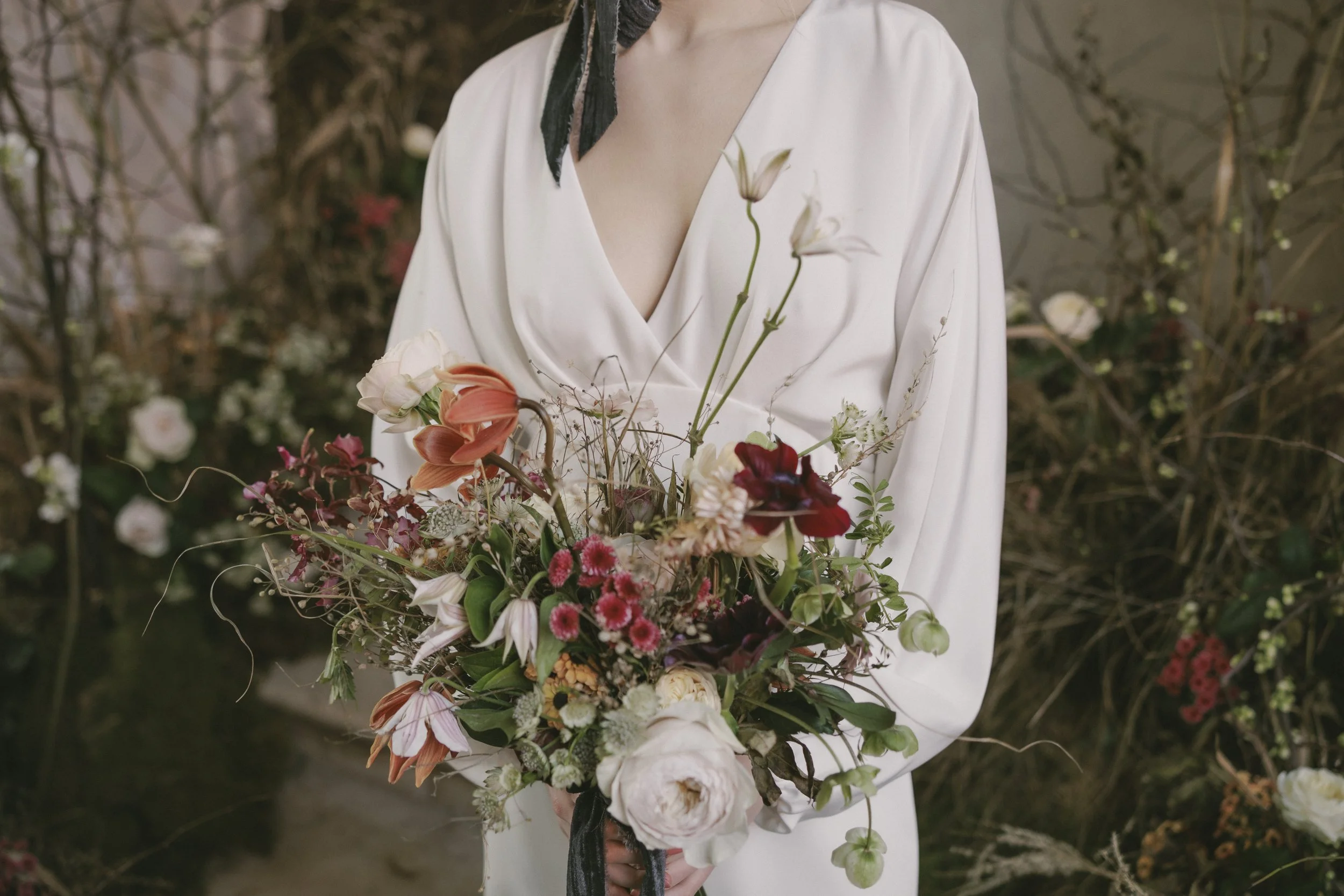A woman in a cream-colored dress holding a bouquet of mixed flowers, including lilies and roses, standing in a garden with a background of bare tree branches and flowers.