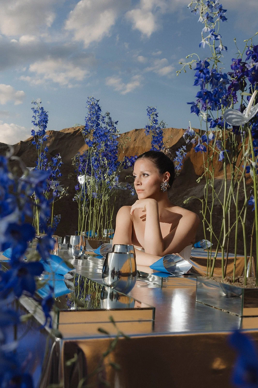 A woman with dark hair in a ponytail, wearing large earrings, sitting at a reflective metallic table surrounded by tall blue flowers. In the background are rocky hills and a partly cloudy sky.