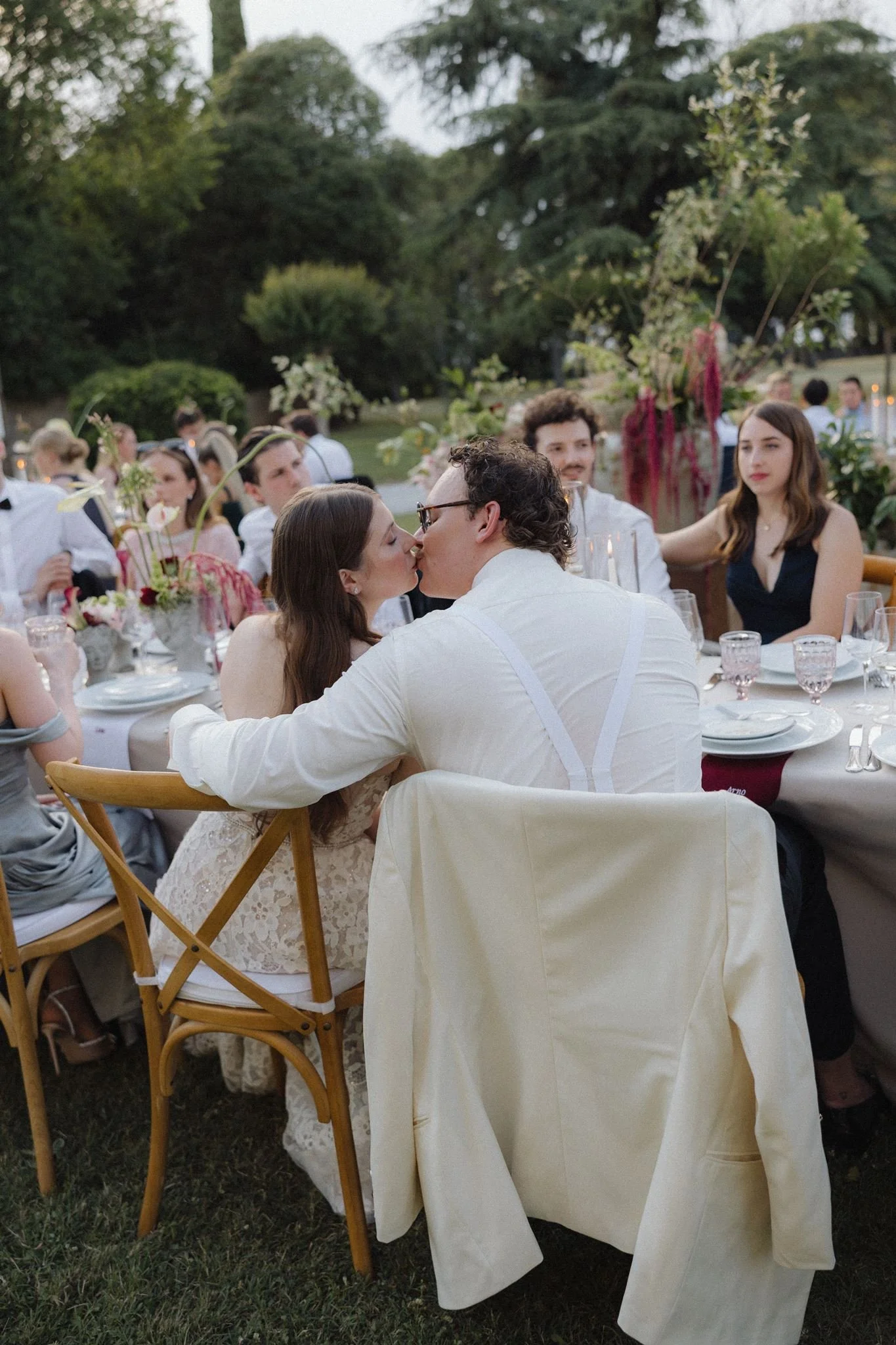 A couple sharing a kiss at an outdoor wedding reception with guests seated at tables with floral centerpieces.