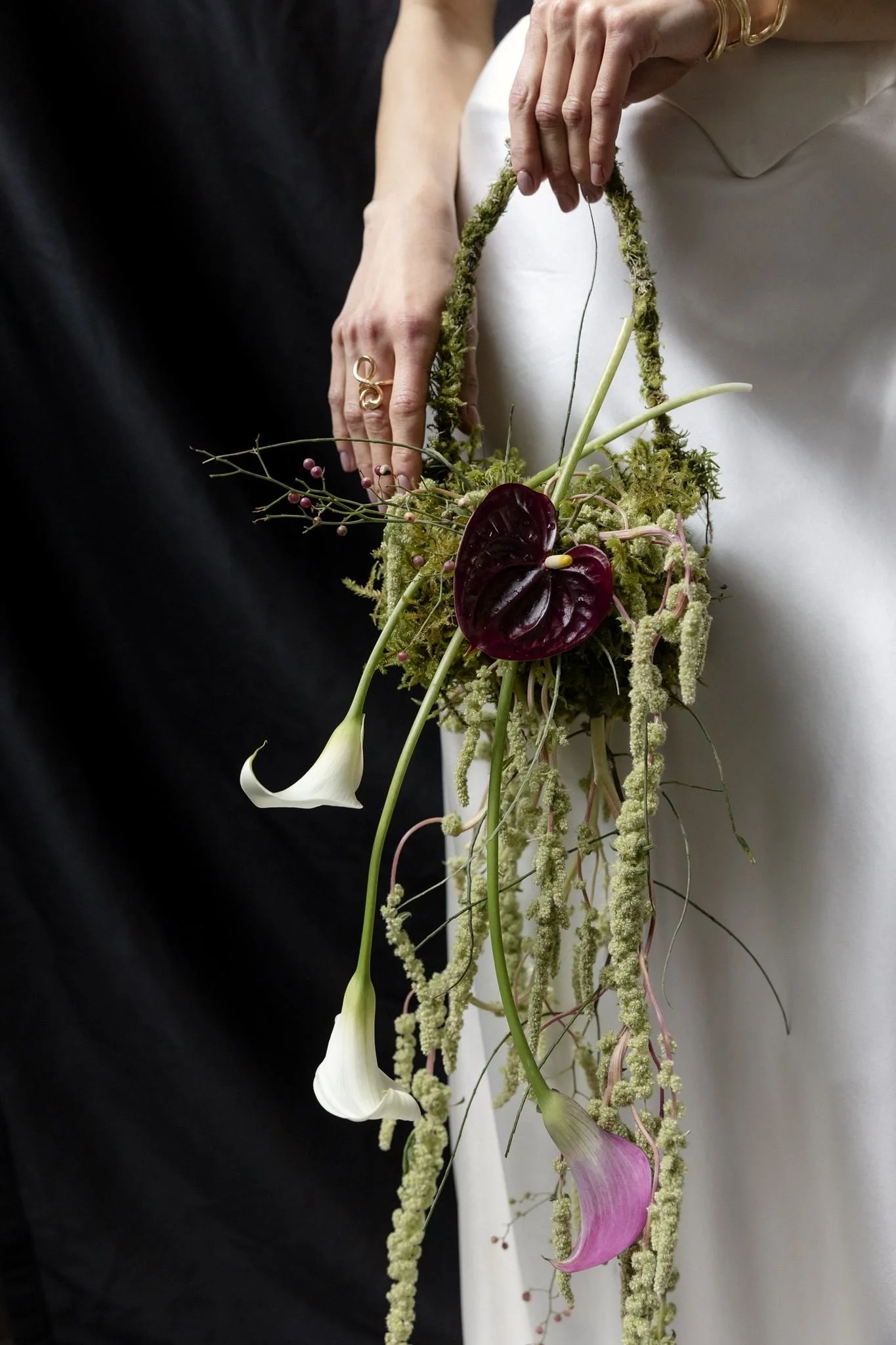 Close-up of a floral arrangement with white calla lilies, pink anthuriums, and various greenery, held by two hands with rings and gold accessories, against a black and white background.