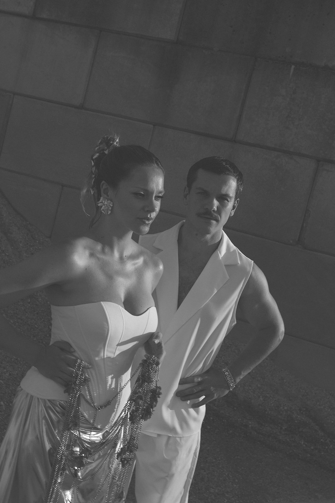 Black and white photo of a woman in a strapless dress with jewelry and a man in a sleeveless vest, both posing against a brick wall.