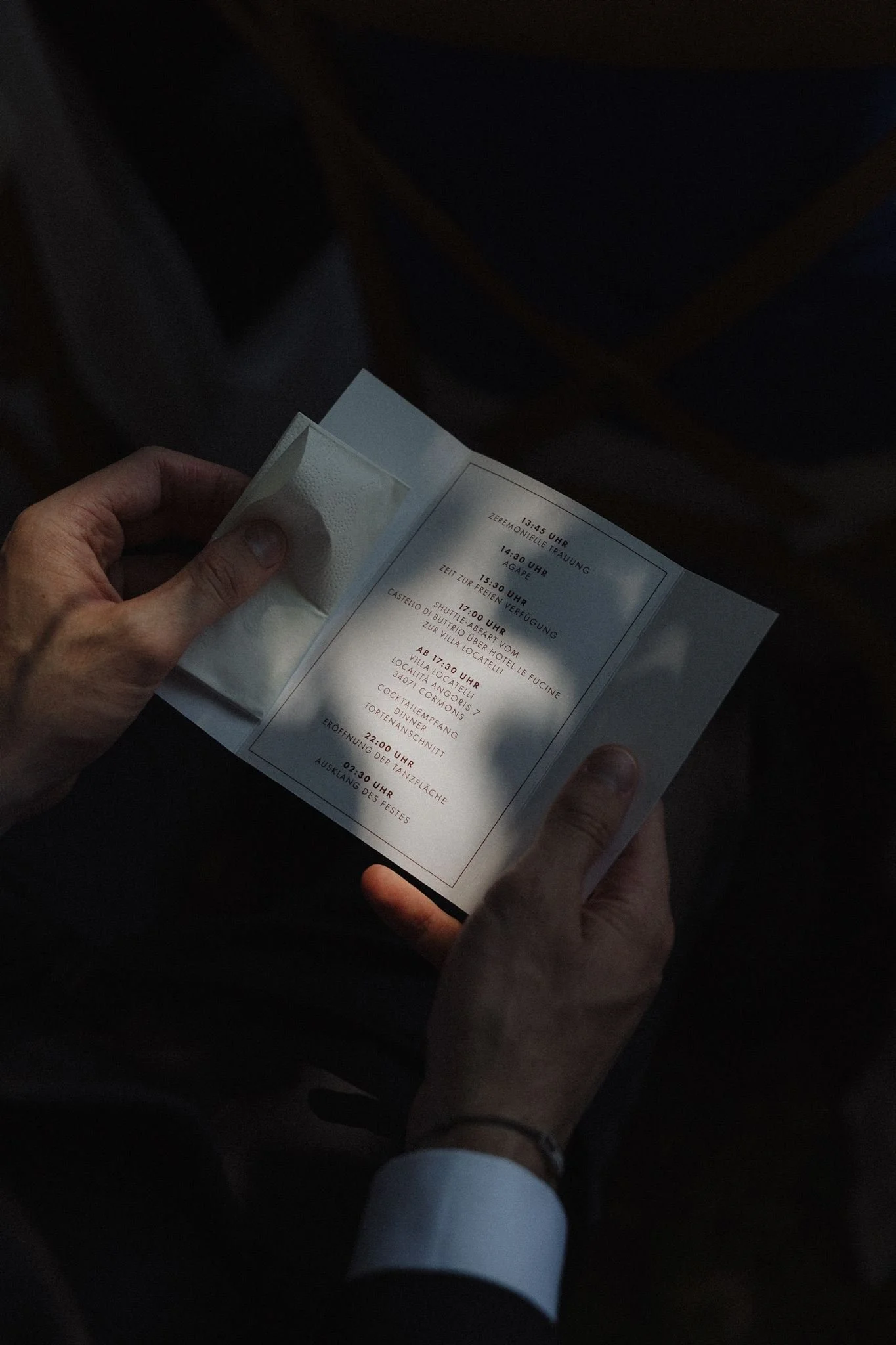 Person holding a wedding itinerary or program, with a dark background and another person's hands visible.