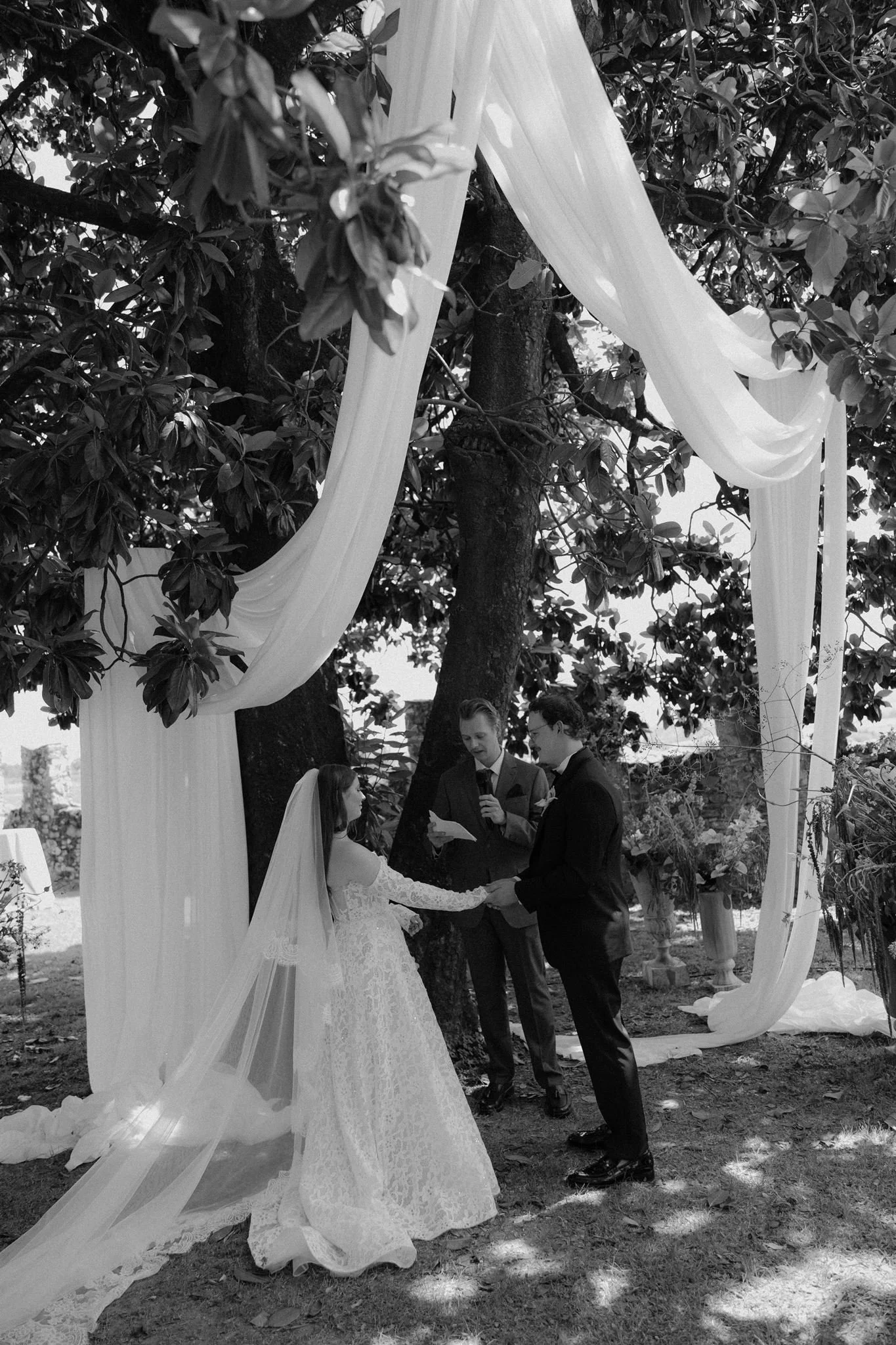 A black and white photo of a wedding ceremony outdoors, with a bride, groom, and officiant standing under fabric drapes and surrounded by trees.