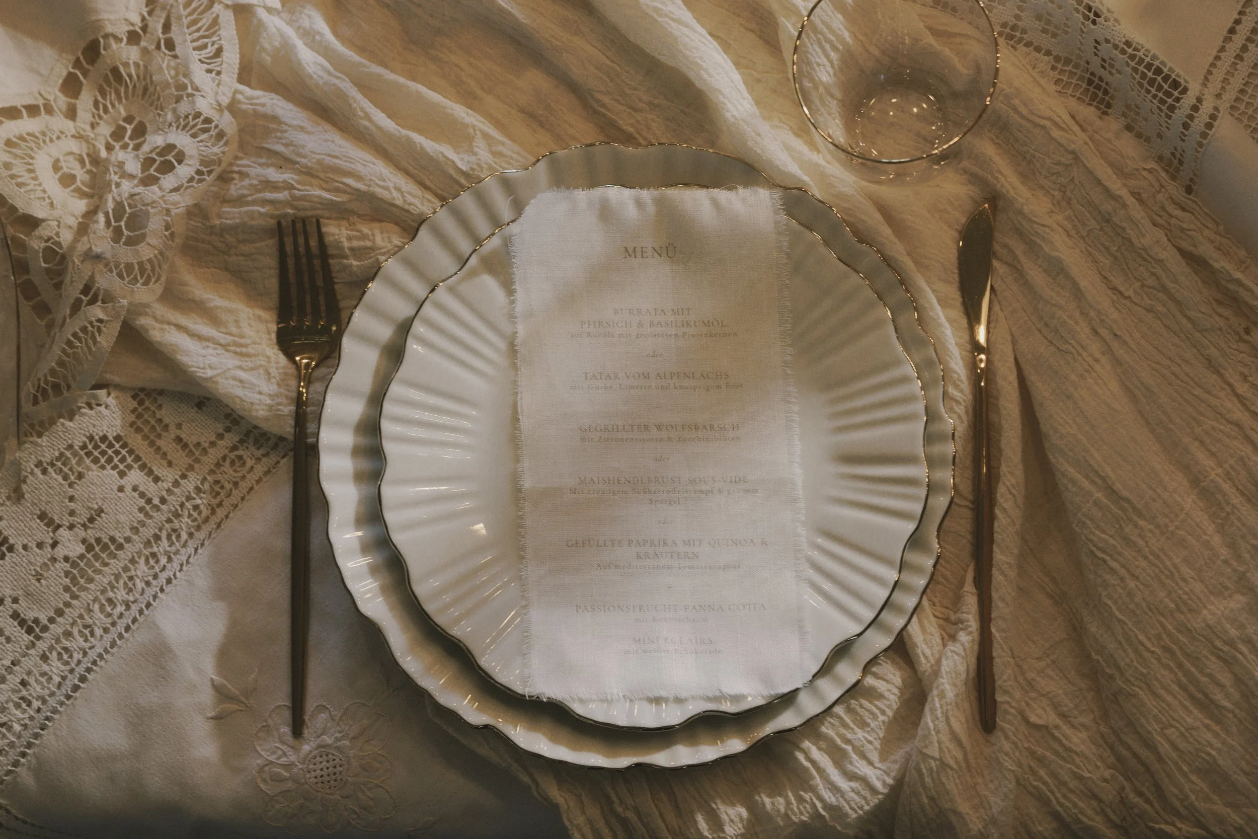 A dining table with lace tablecloth, a decorative doily, and a large, scalloped-edge plate with a menu inside. There is a glass and cutlery aligned on each side of the plate.