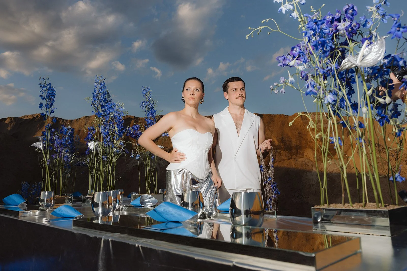 Two models in white outfits standing outdoors among tall blue flowers, with a rocky landscape and cloudy sky in the background.