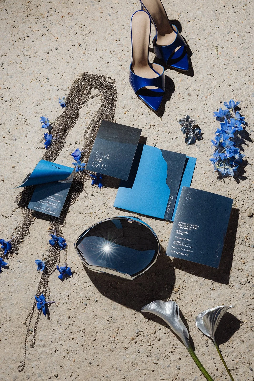 A collection of accessories and items on sandy ground, including a pair of blue high-heeled shoes, blue flowers, a silver necklace, a pair of earrings, a black bowl, two silver shell-shaped objects, and blue and black printed cards.