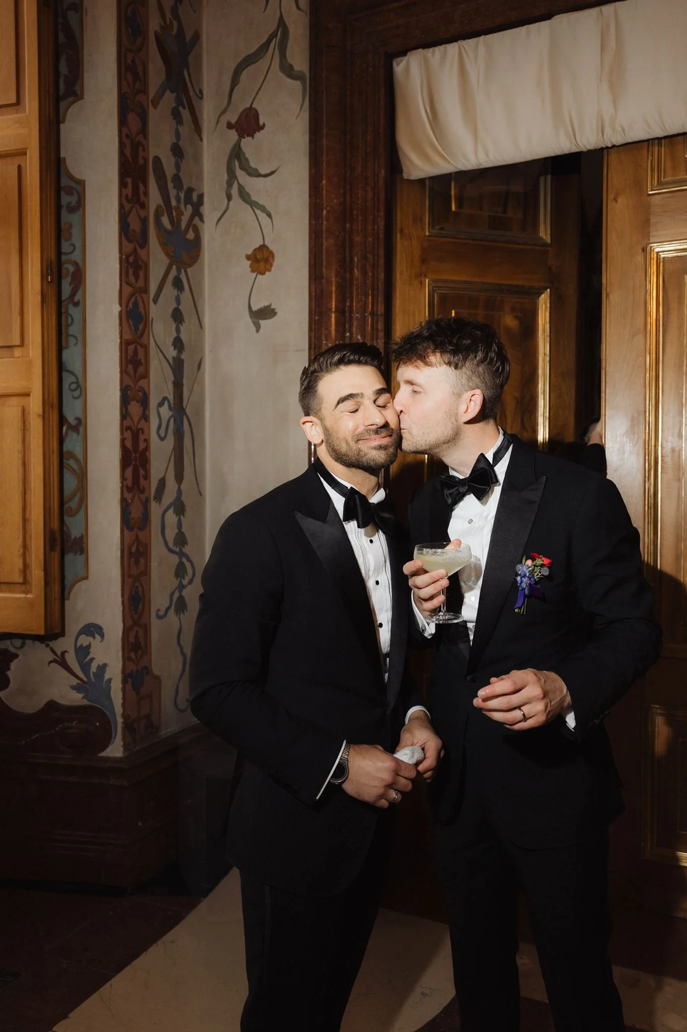Two men in tuxedos sharing a kiss at a formal event, with one holding a cocktail and the other holding a towel.