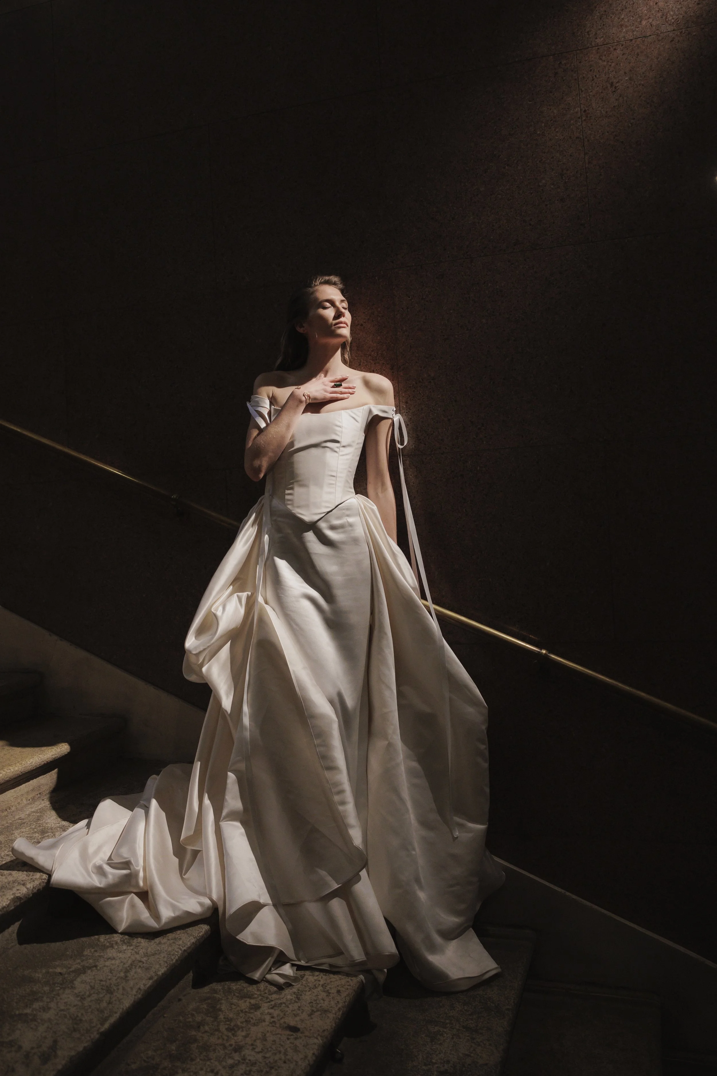 A woman standing on a stone staircase wearing an elegant off-the-shoulder white gown, with her eyes closed and hand on her chest, illuminated by a soft light against a dark wall background.