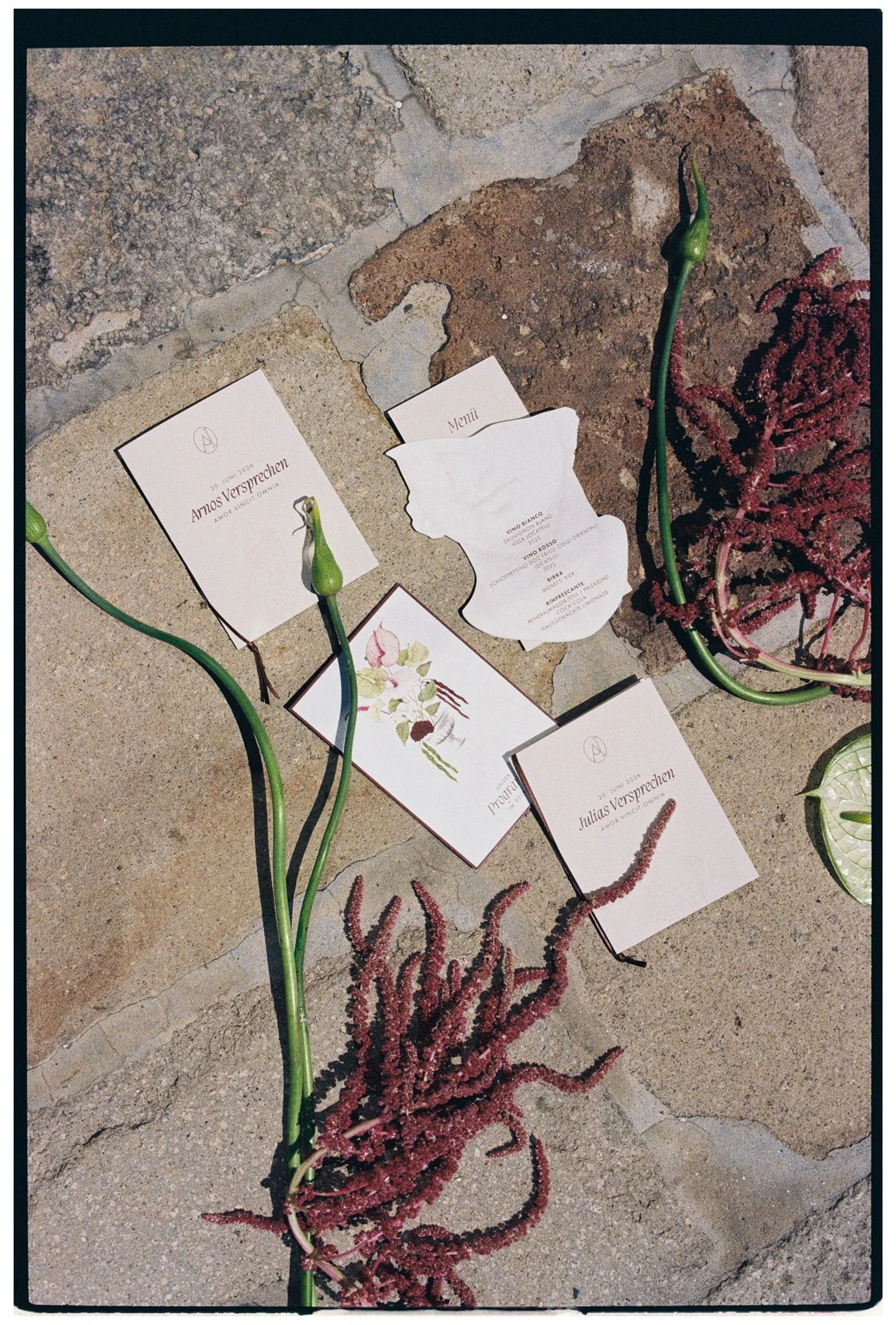 Several menus and event programs laid out on a concrete and stone surface, with long green stems, pink, red and purple flower arrangements, and decorative red and pink floral designs.