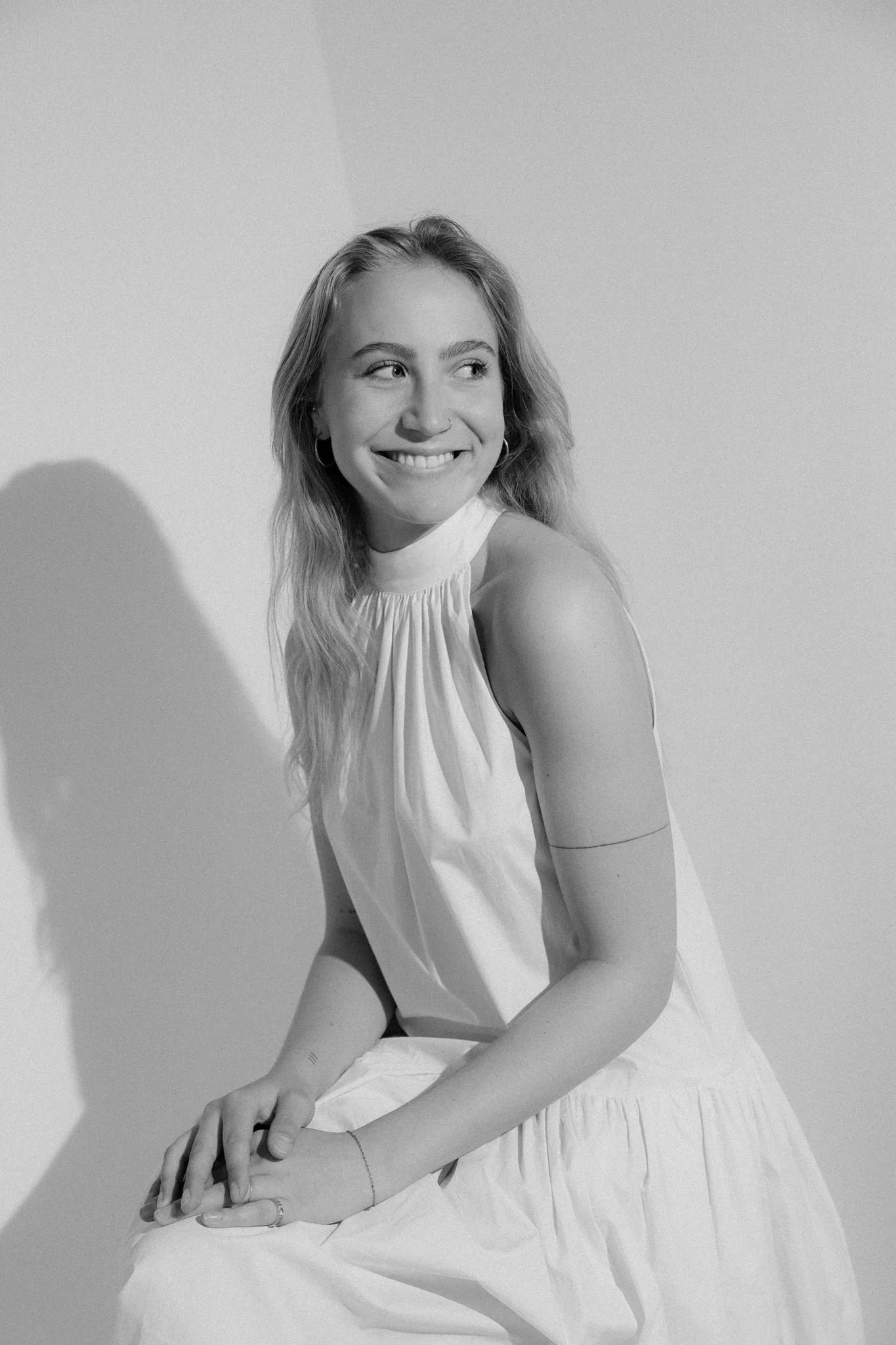 A smiling woman with long hair and hoop earrings, sitting against a plain background, wearing a sleeveless top, in black and white photograph.